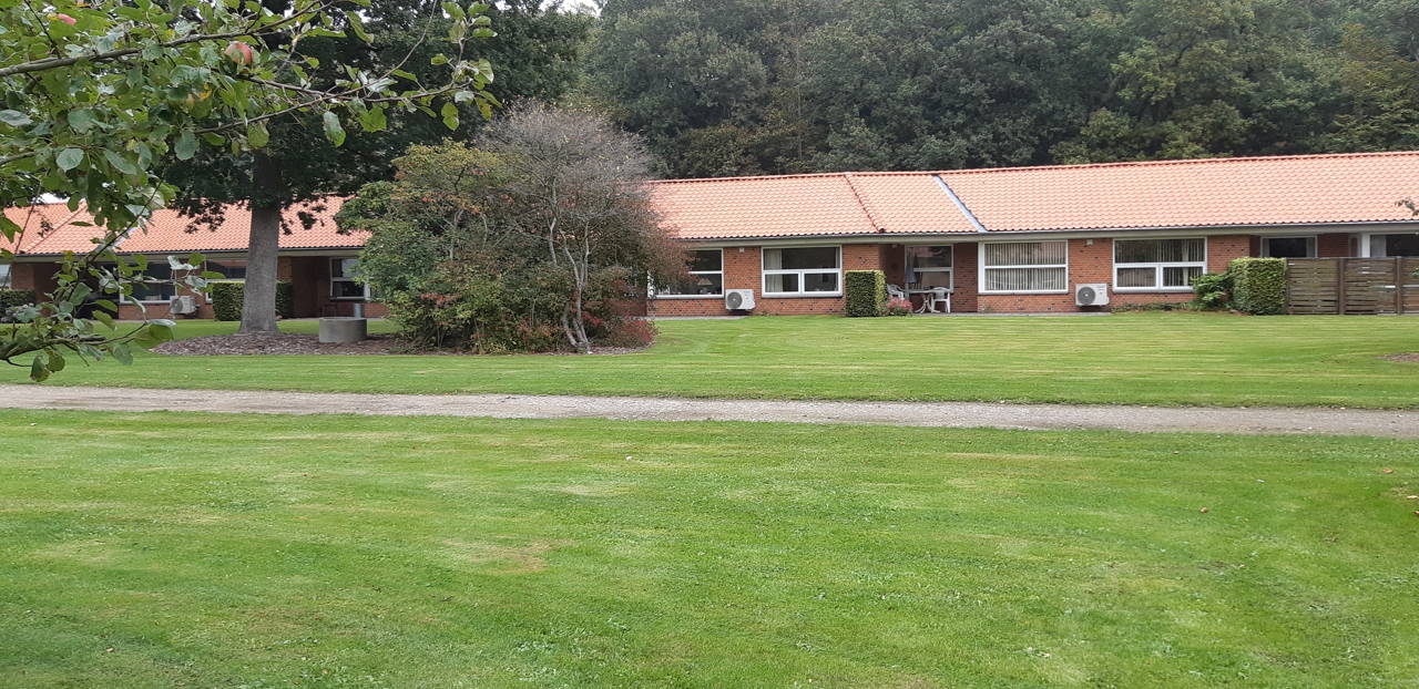 Solgården set ude fra, bygningen er en lang og der er røde mursten og hvide vinduer. Der er en stor flot grøn græsplæne og to store træer.