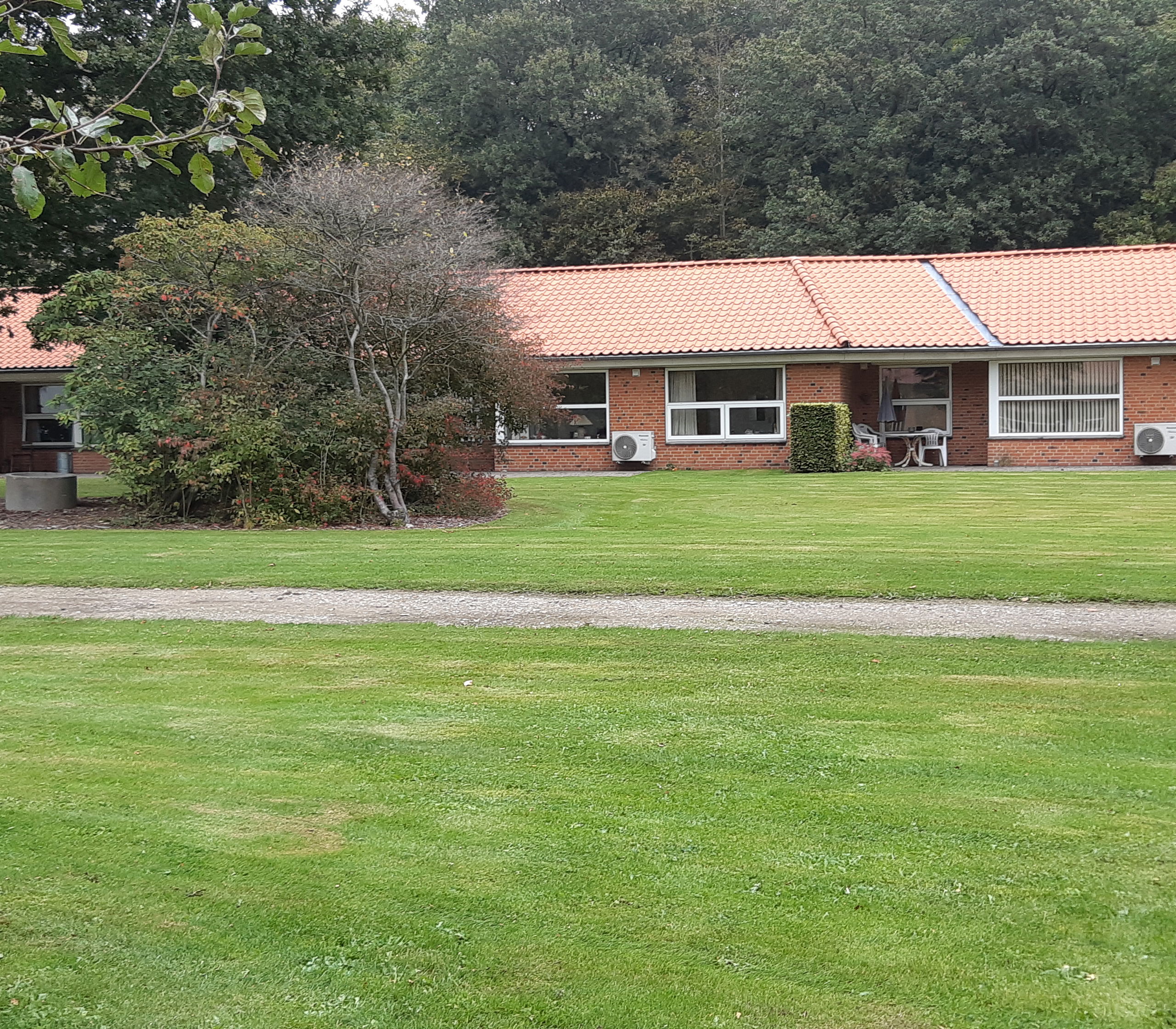 Solgården set ude fra, bygningen er en lang og der er røde mursten og hvide vinduer. Der er en stor flot grøn græsplæne og to store træer.