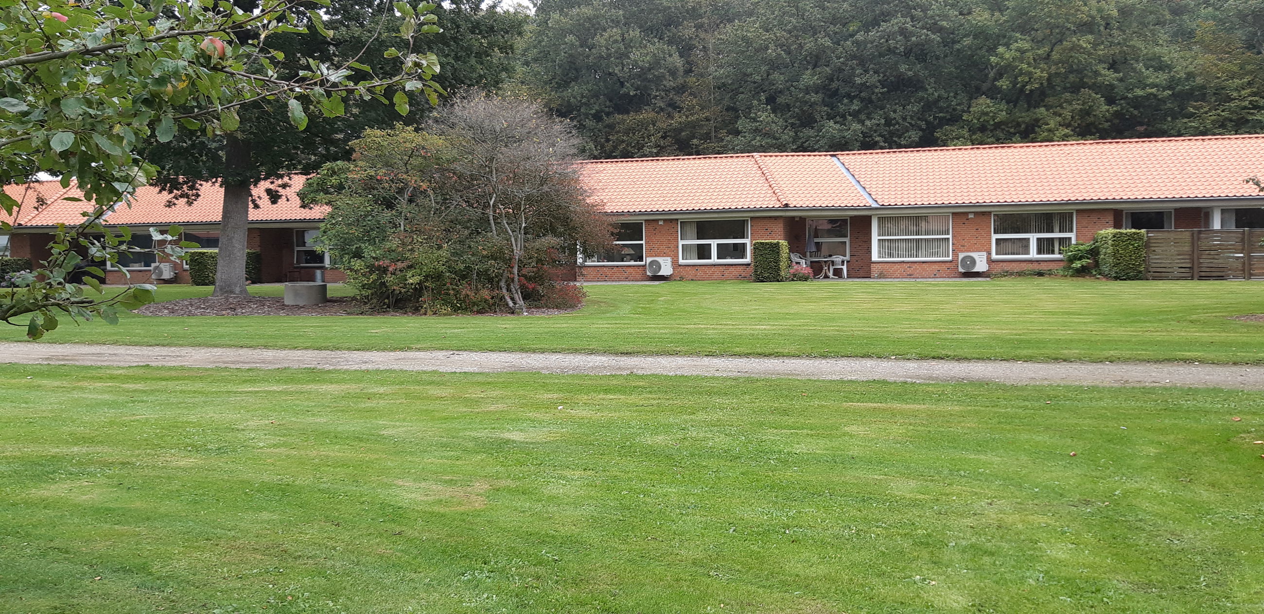 Solgården set ude fra, bygningen er en lang og der er røde mursten og hvide vinduer. Der er en stor flot grøn græsplæne og to store træer.