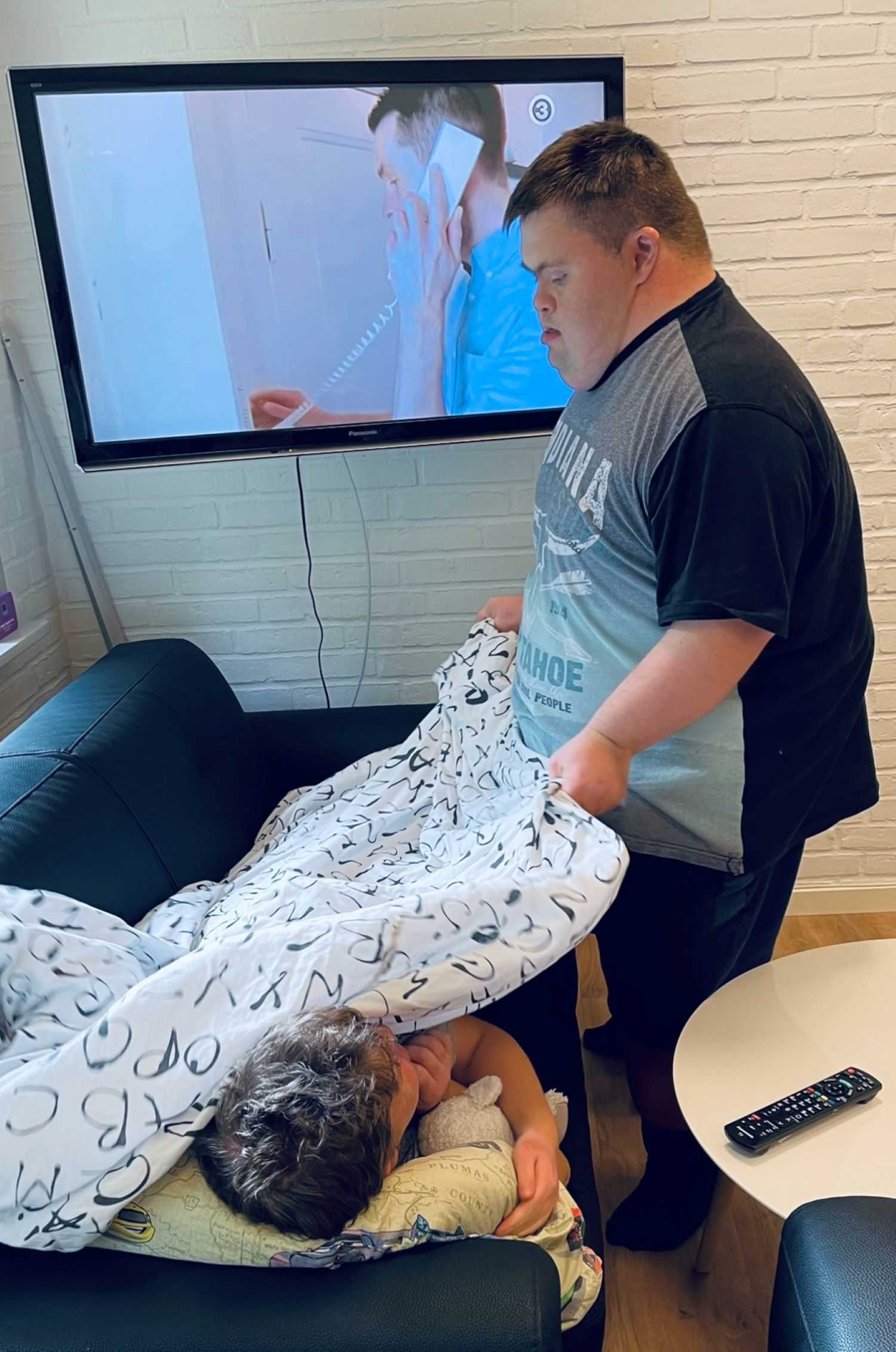 En borger putter dyne på en anden borger i en sofa, bag dem på væggen er stort fladskærmstv.