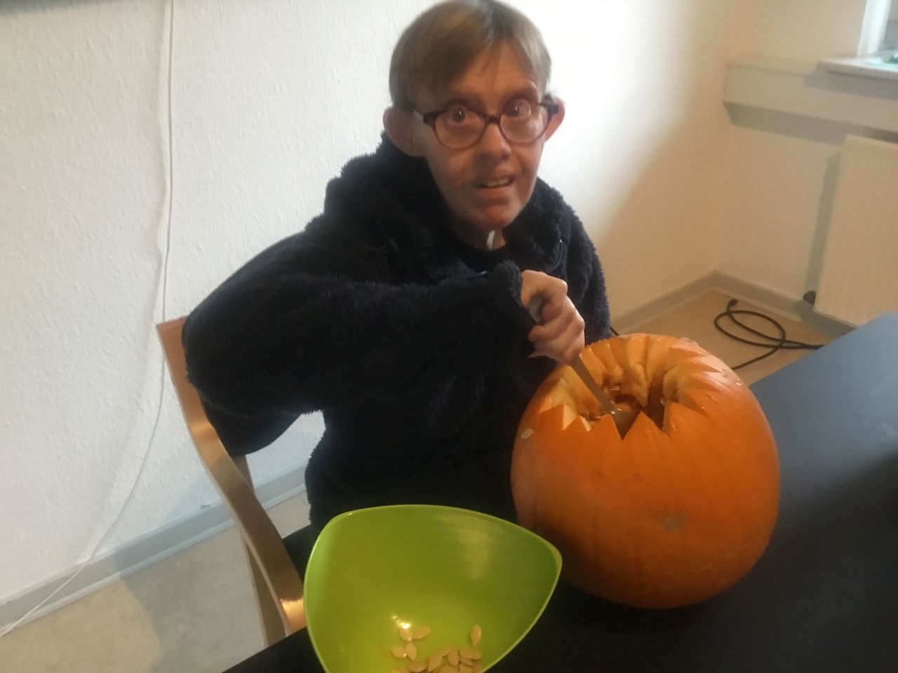 En borger er ved at lave et græskarhoved af et stort græskar, man kan se at han tager kerner ud og putter i en grøn skål.