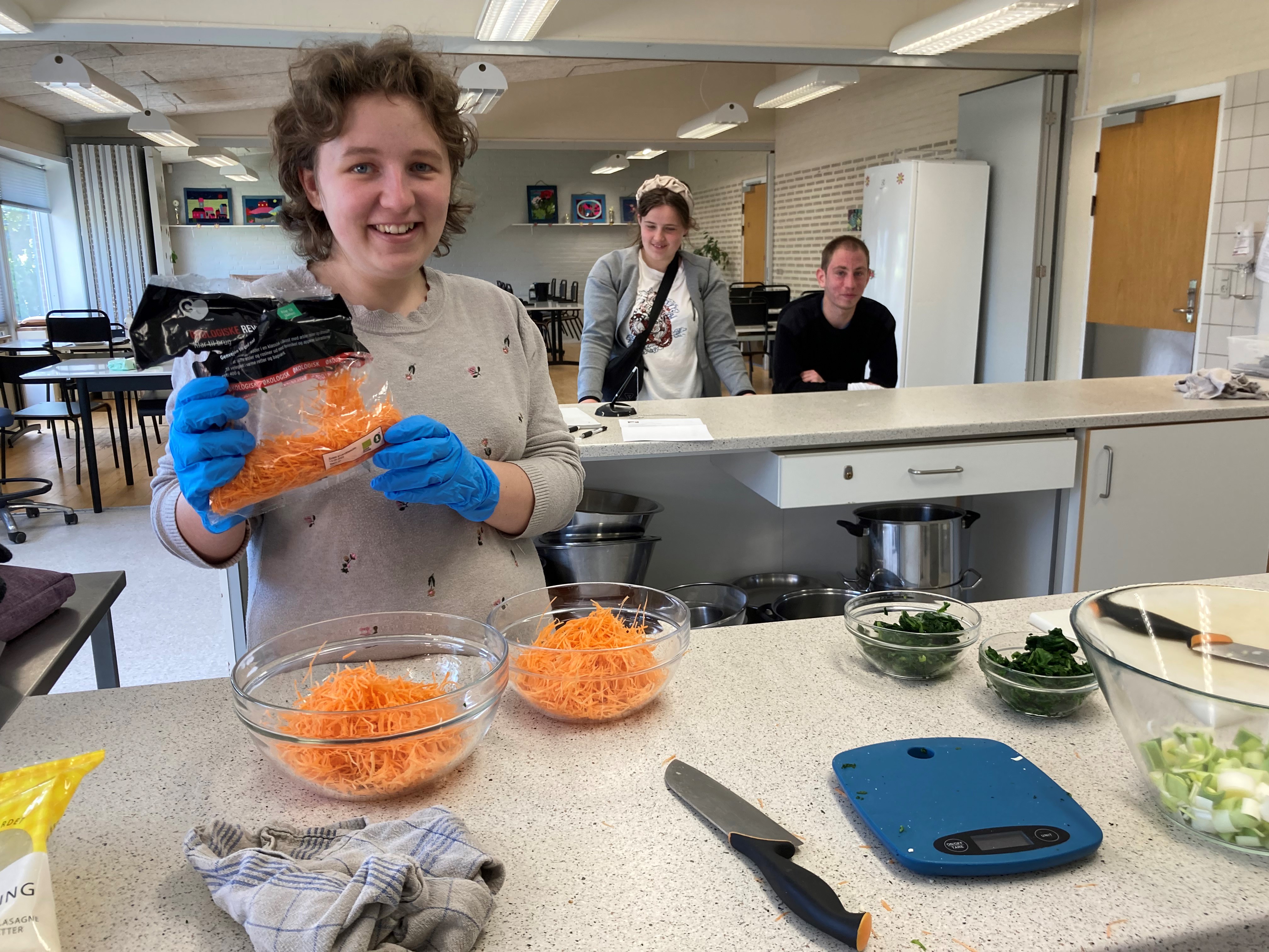 En borger er ved at putte revet gulerod i to glasskåle, bag hende står to andre borgere og kigger på.