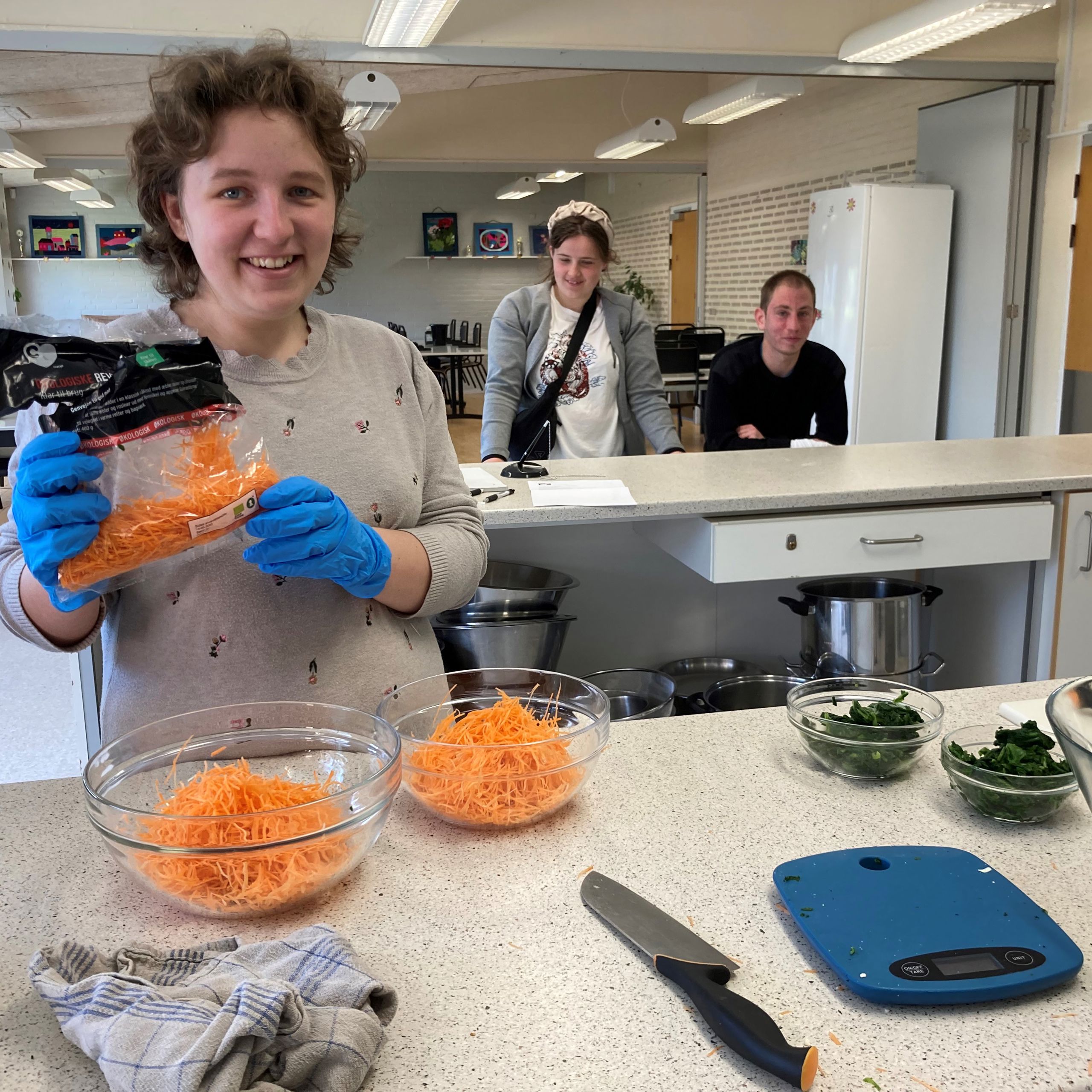 En borger er ved at putte revet gulerod i to glasskåle, bag hende står to andre borgere og kigger på.