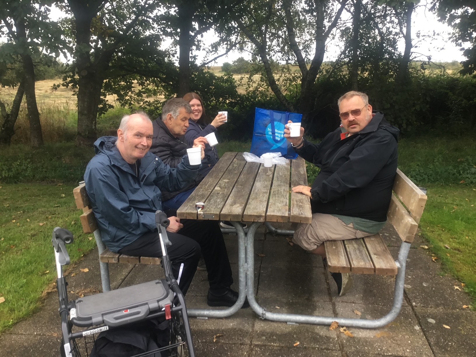 Fire borgere skåler i kaffe ved et bord og bænkesæt, det ligner det lige har regnet  fliserne under dem er våde.