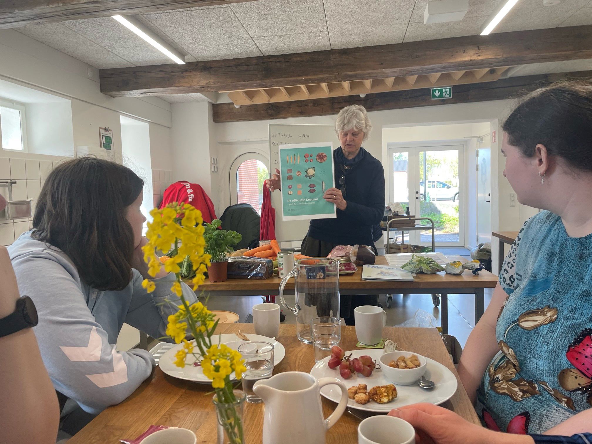 To kvinder sidder ved et bord og lytter til en anden kvinde der giver et foredrag om Højergaard. På bordet hvor kvinderne sidder står der en vase med smukke gule blomster i. Foran det bord er der placeret et andet bord hvor kvinden der holder foredraget står. Hun står med en plakat med fødevarer på i hånden.