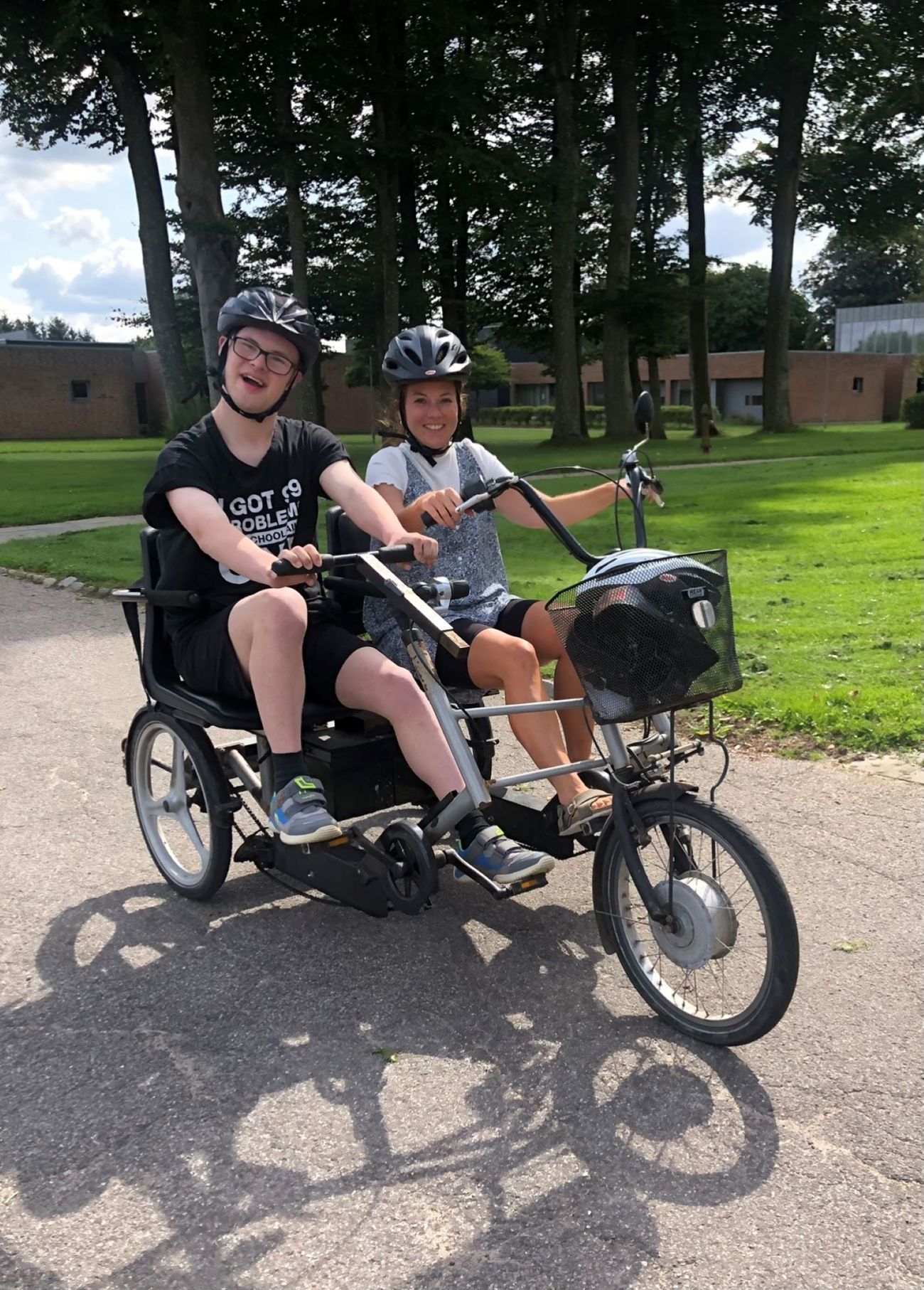 En borger på cykel med en hjælper de har begge cykelhjelm på, bag des ses bygninger på løgumgårde samt græsplæner og stor træer.