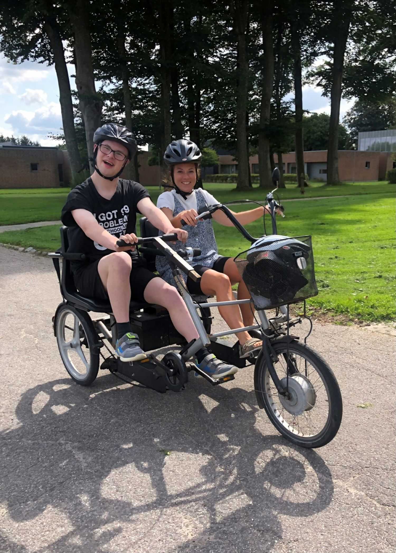 En borger på cykel med en hjælper de har begge cykelhjelm på, bag des ses bygninger på løgumgårde samt græsplæner og stor træer.