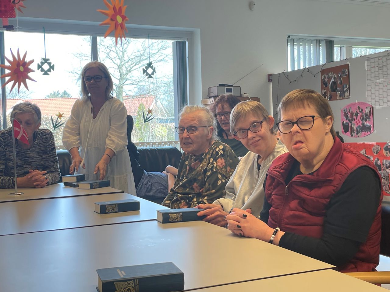Nogle borgere sidder rundt om et bord foran dem ligger en blå sangbog fra folkehøjskolen, nederst på bordet står et flag foran en borger, ned fra loftet hænger der orange blomster