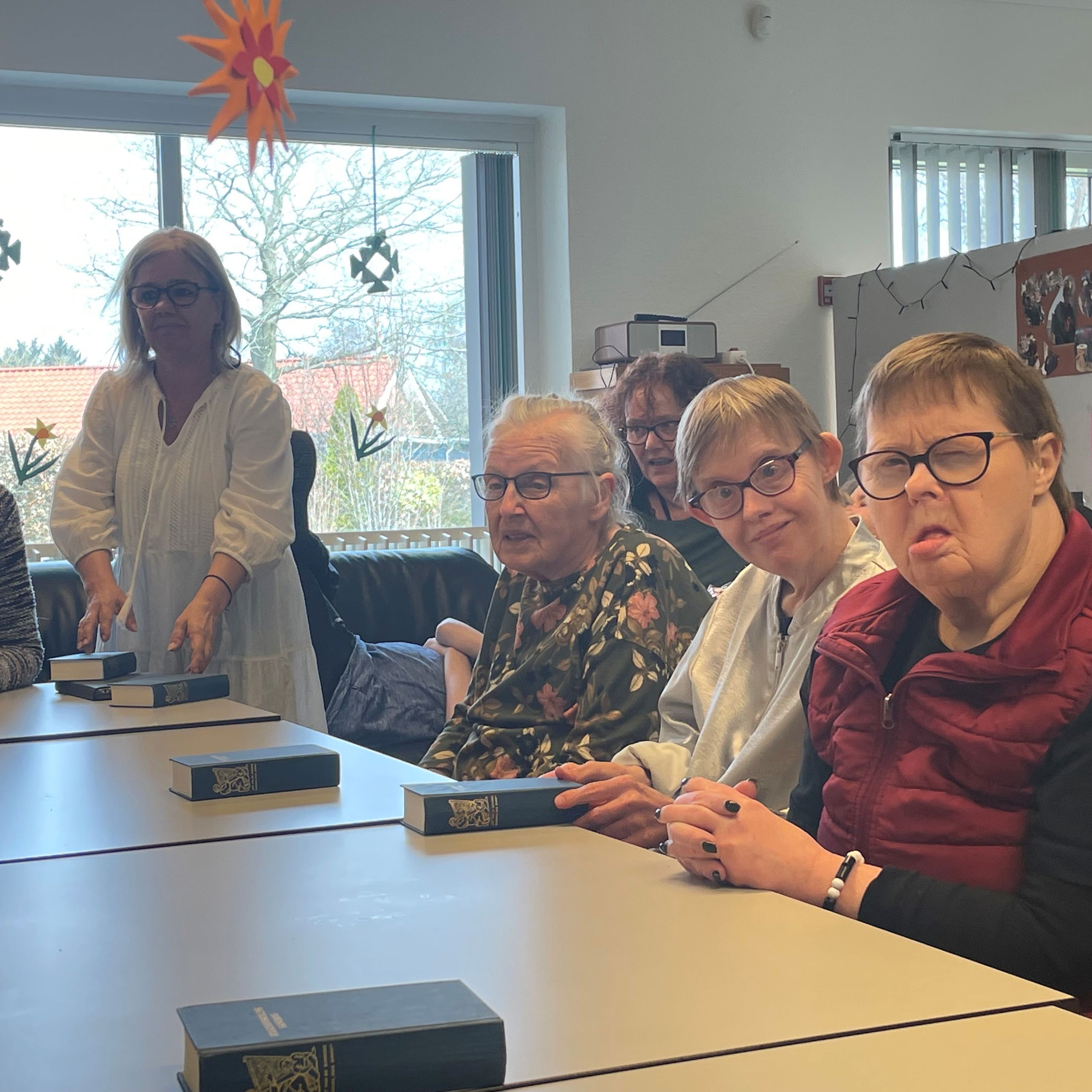 Nogle borgere sidder rundt om et bord foran dem ligger en blå sangbog fra folkehøjskolen, nederst på bordet står et flag foran en borger, ned fra loftet hænger der orange blomster