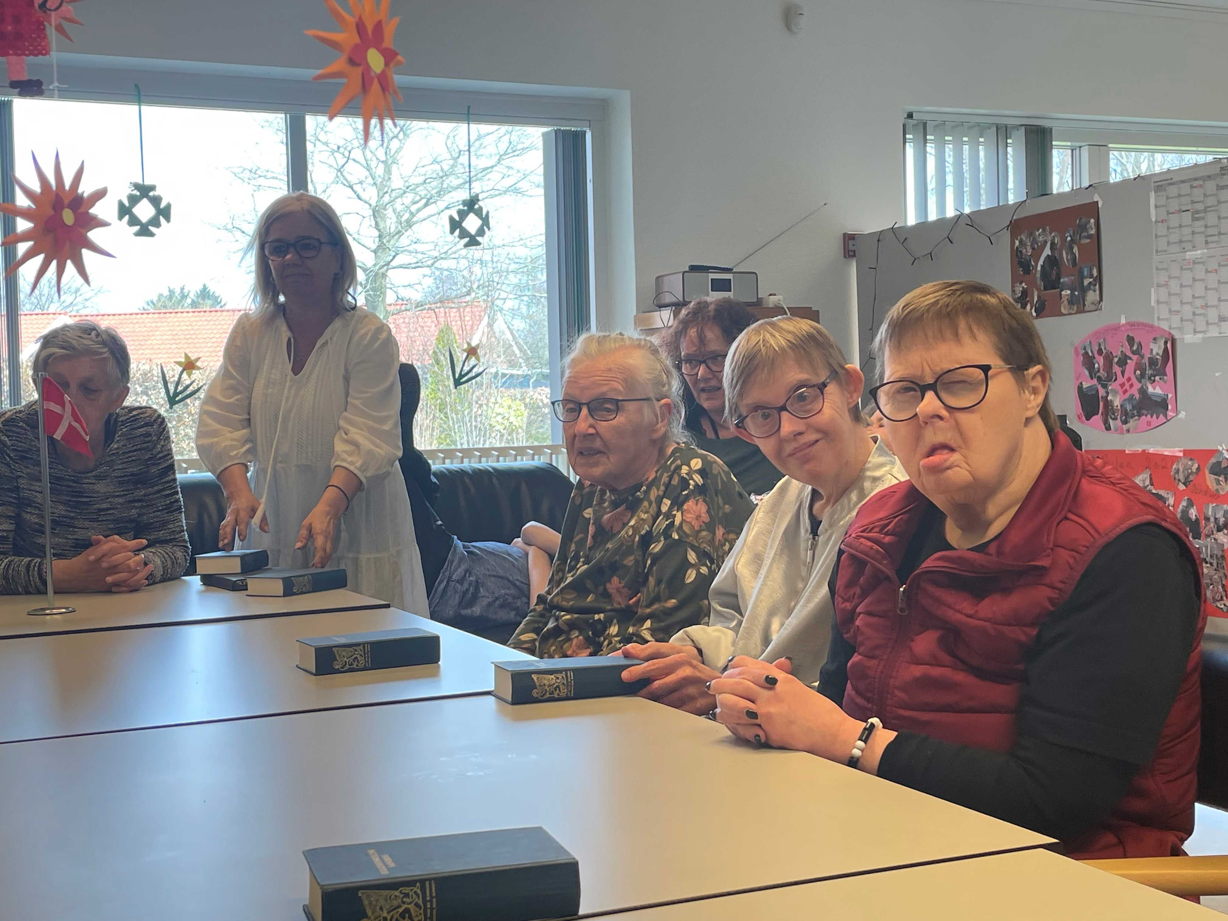 Nogle borgere sidder rundt om et bord foran dem ligger en blå sangbog fra folkehøjskolen, nederst på bordet står et flag foran en borger, ned fra loftet hænger der orange blomster