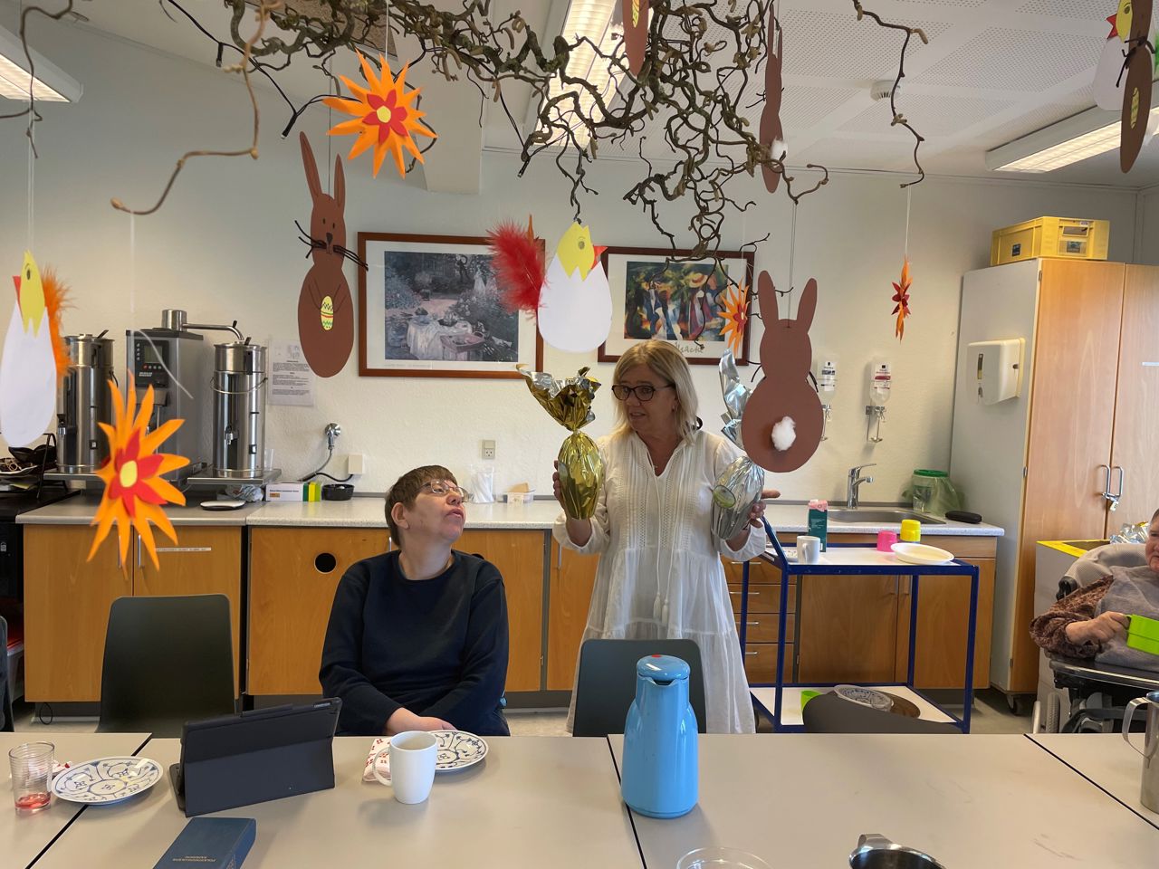 To borgere sidder ved et bord mens en ansat står med to påskeæg og er ved at fortælle noget, i loftet hænger udklippet påskepynt på en troldhasselgren