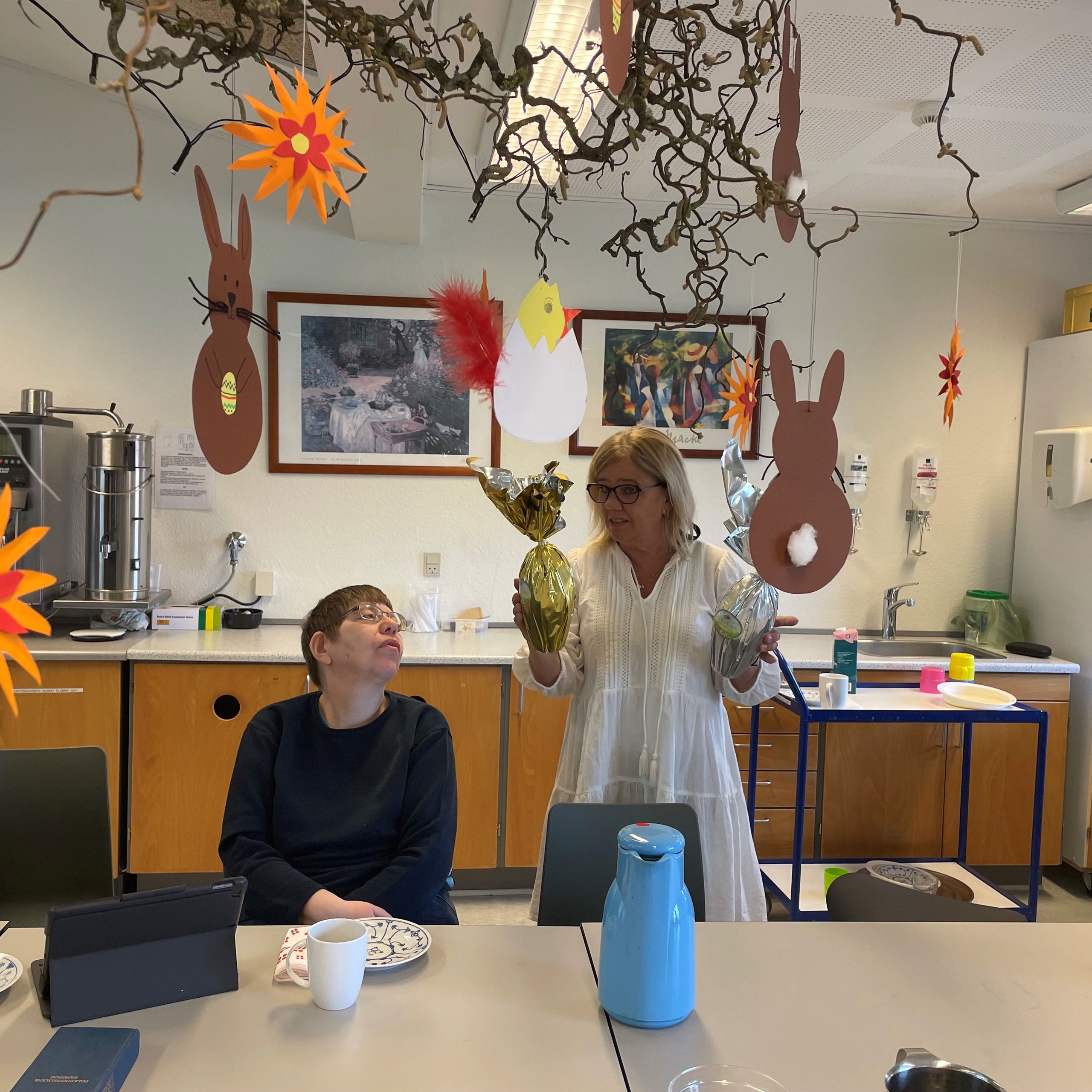 To borgere sidder ved et bord mens en ansat står med to påskeæg og er ved at fortælle noget, i loftet hænger udklippet påskepynt på en troldhasselgren