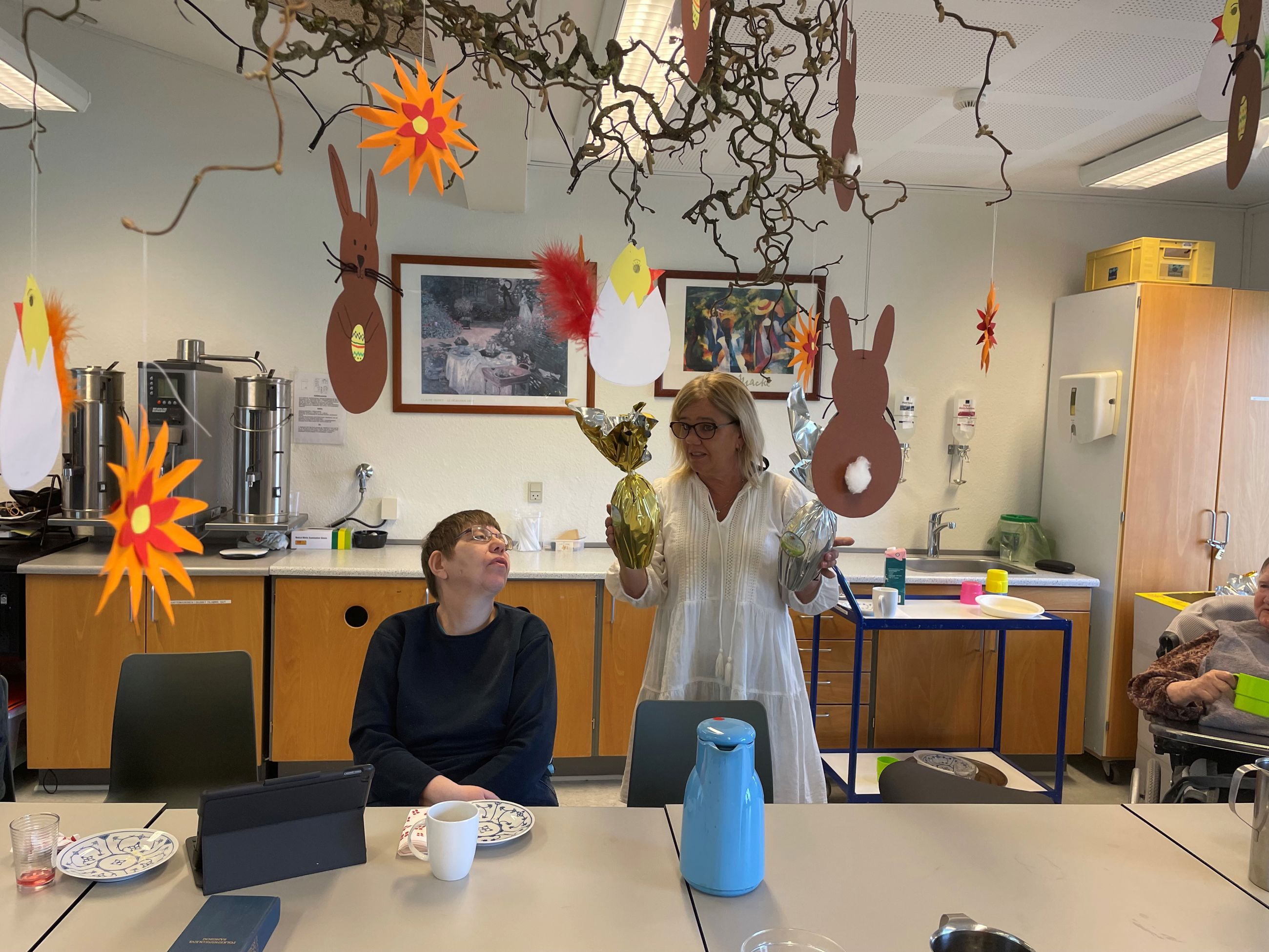 To borgere sidder ved et bord mens en ansat står med to påskeæg og er ved at fortælle noget, i loftet hænger udklippet påskepynt på en troldhasselgren