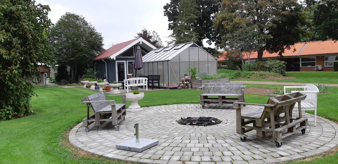 Bålplads hvor der er tre bænke lavet af træpaller og i baggrunden er der et hus med et drivhus foran.