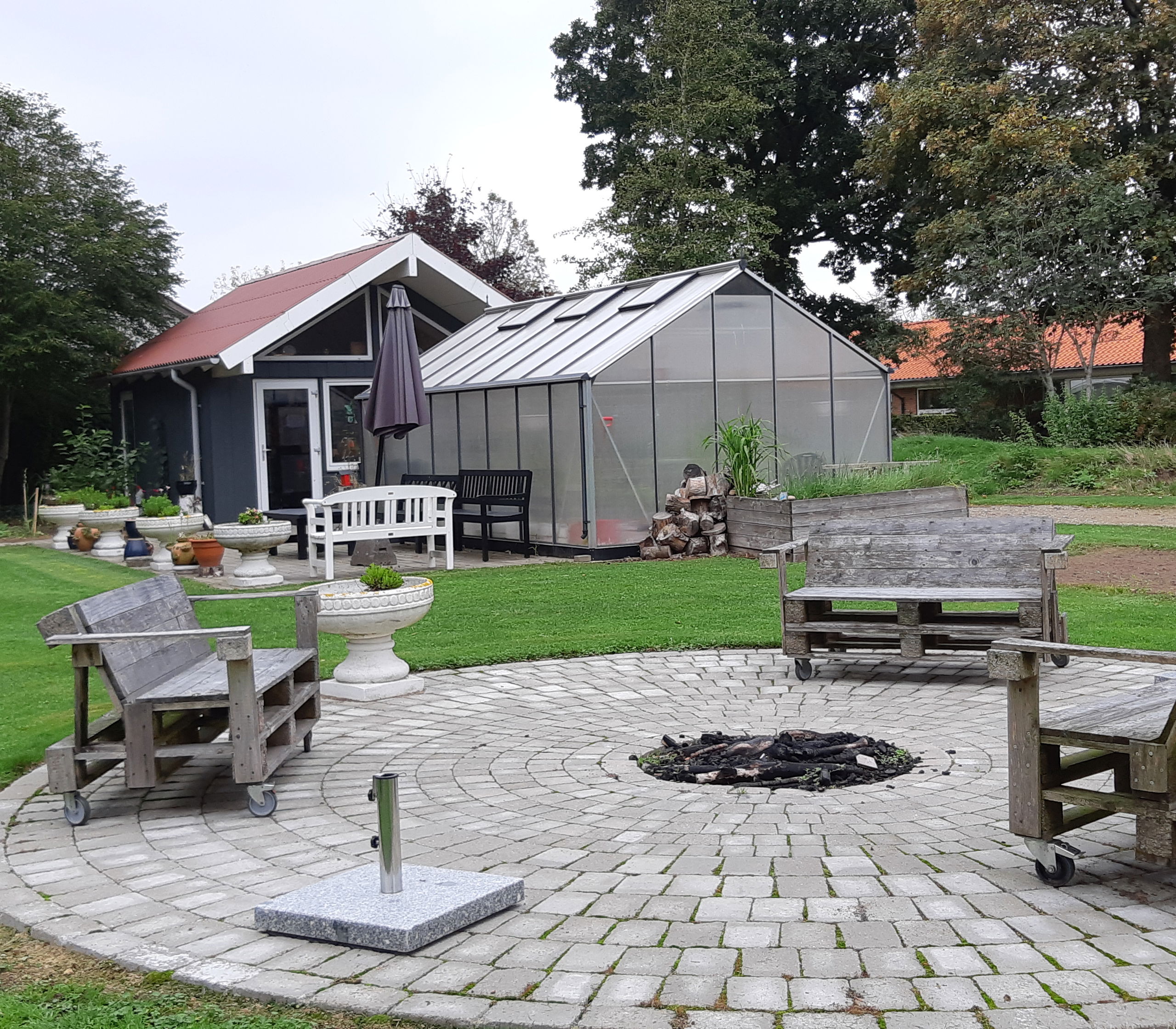 Bålplads hvor der er tre bænke lavet af træpaller og i baggrunden er der et hus med et drivhus foran.
