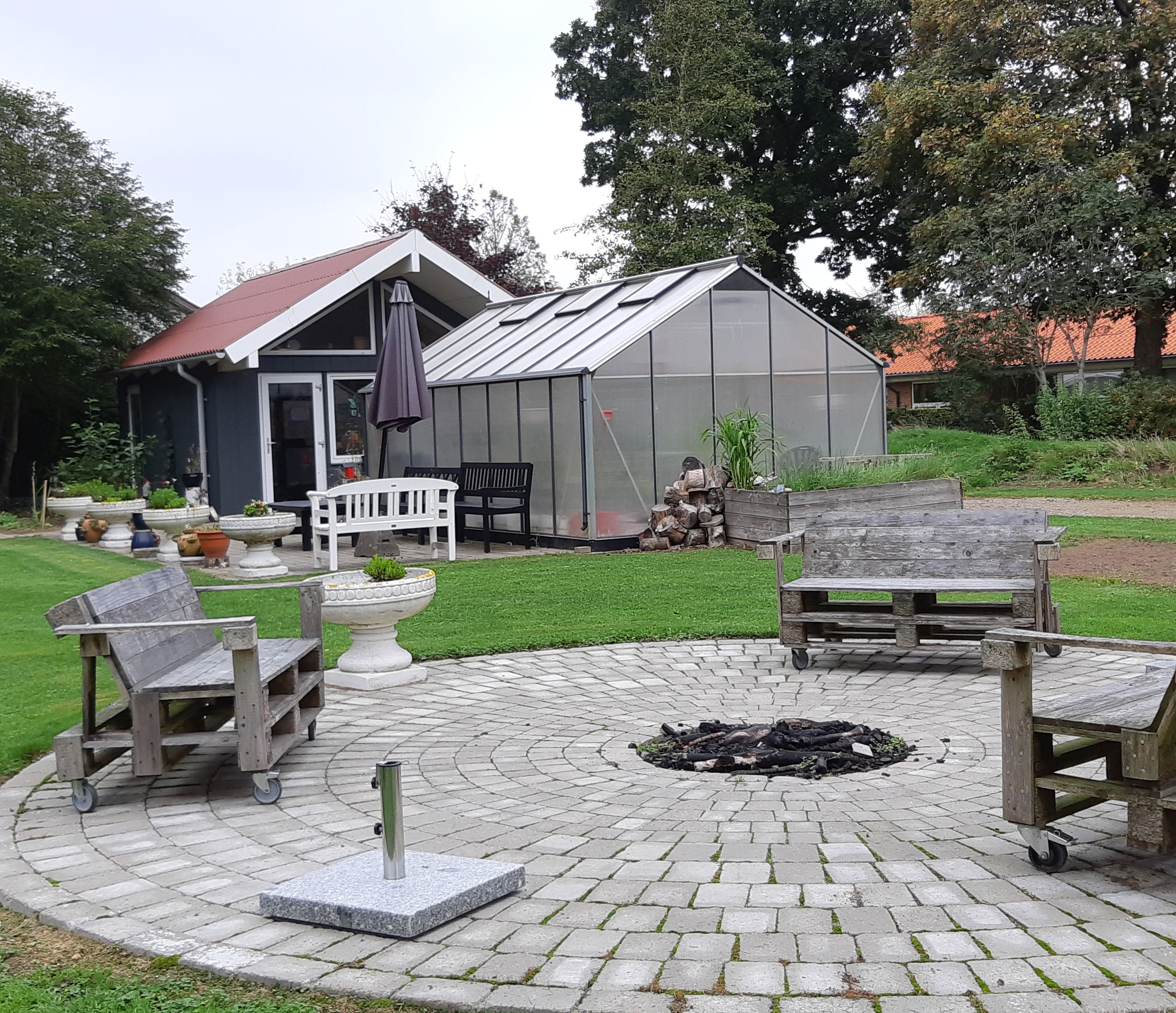 Bålplads hvor der er tre bænke lavet af træpaller og i baggrunden er der et hus med et drivhus foran.