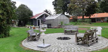 Bålplads hvor der er tre bænke lavet af træpaller og i baggrunden er der et hus med et drivhus foran.