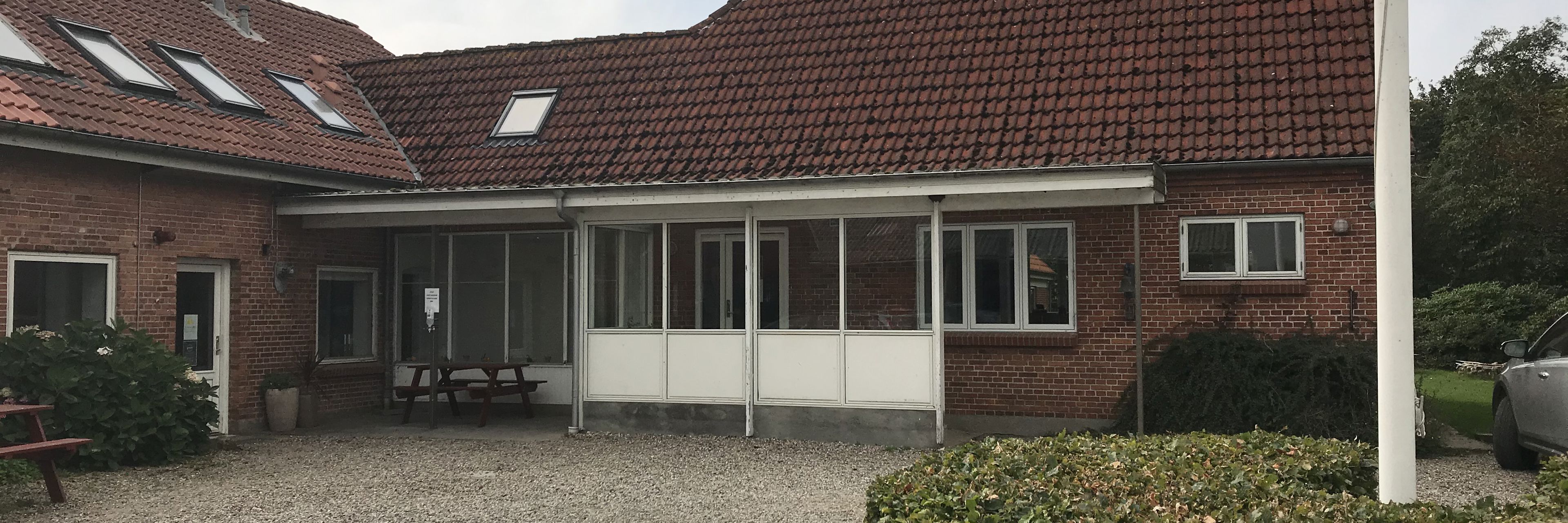 hovedbygning i røde sten og rød tegltag, indgang med glasparti foran, en gårdsplads med grus og i midten står der en flagstang og rundt om den en lav grøn hæk.