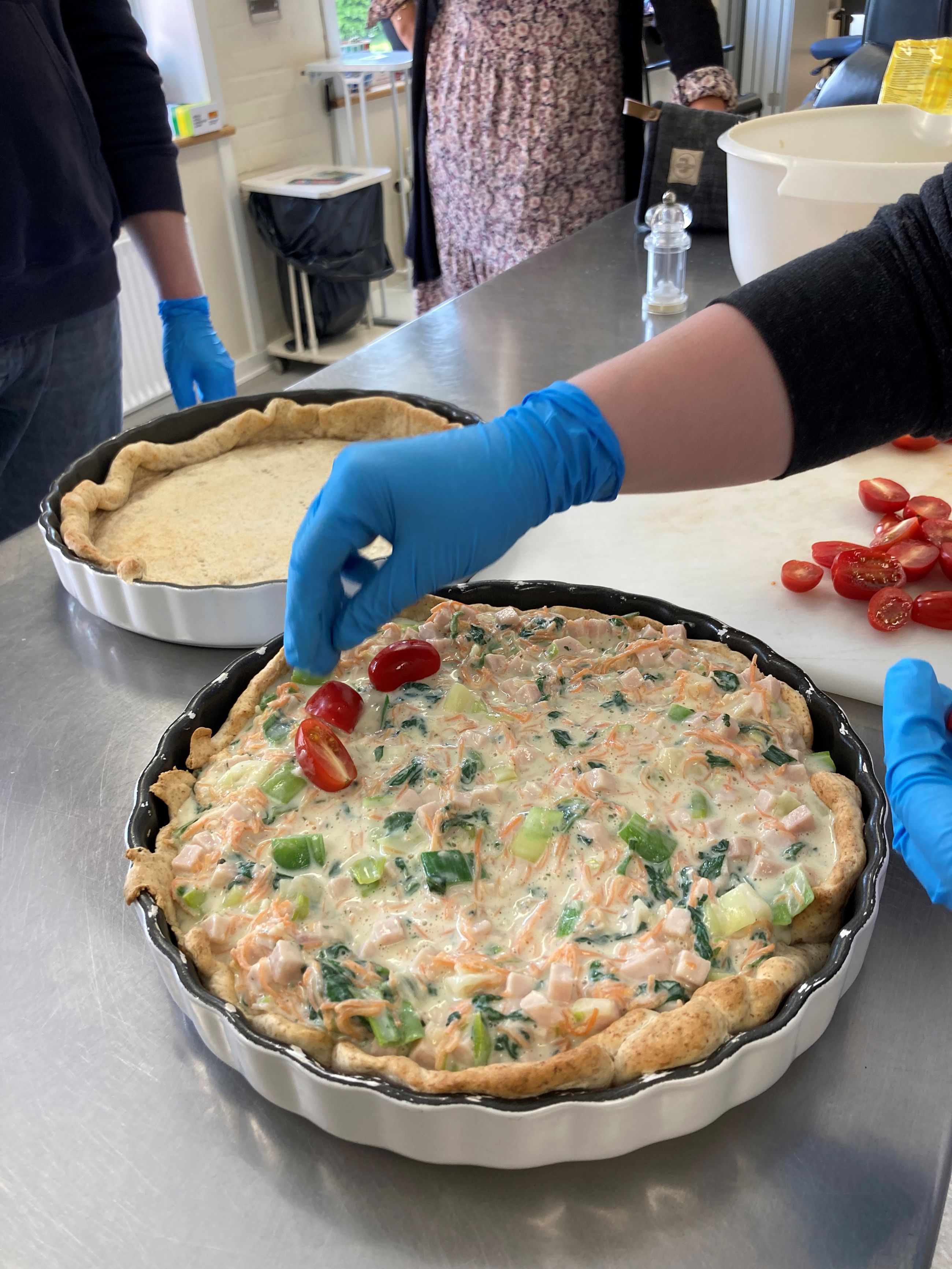 Tomaterne ligges ovenpå tærten ved siden af ses en tæret uden fyld, endnu bagved står andre borgere og kigger på.