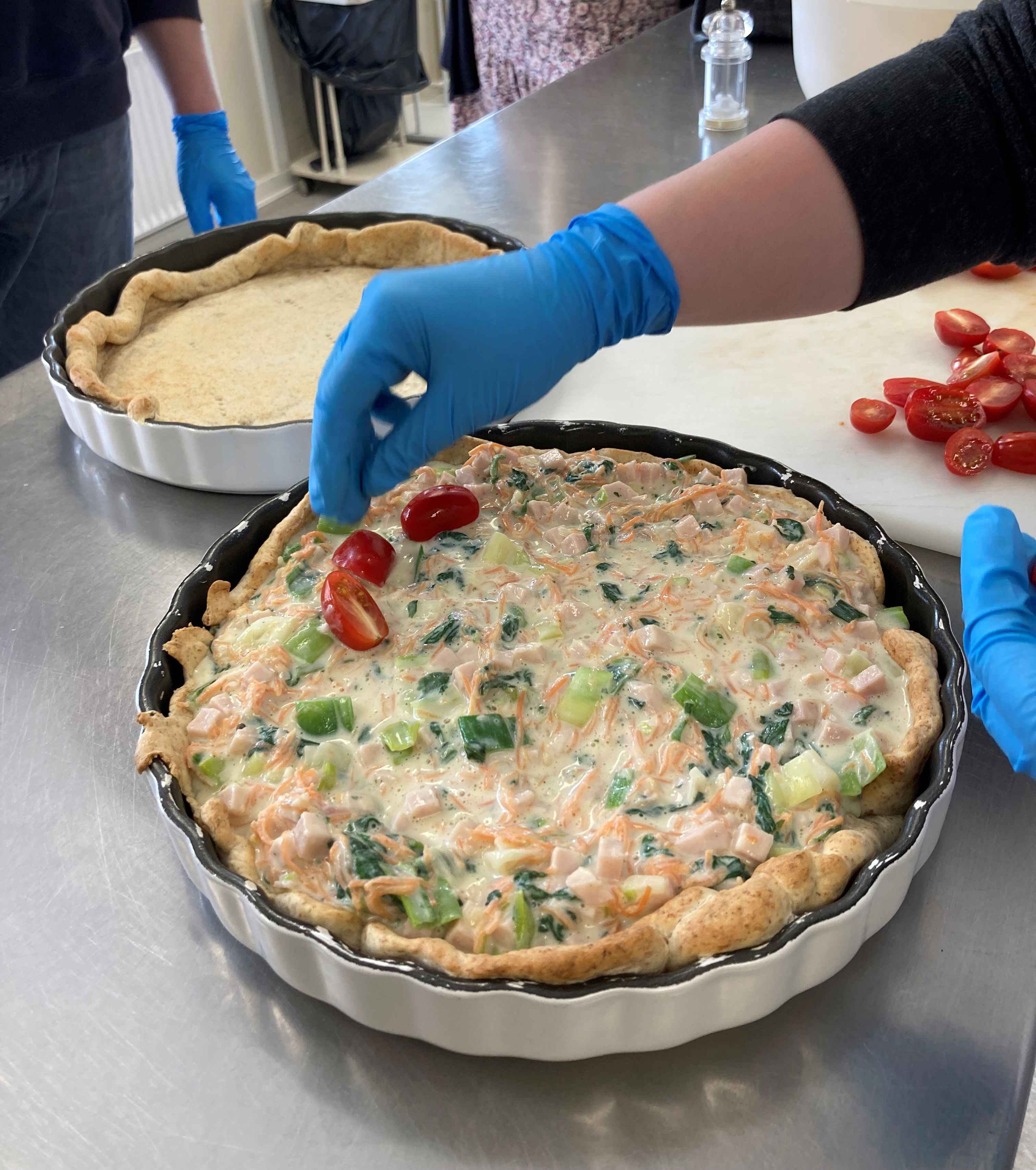 Tomaterne ligges ovenpå tærten ved siden af ses en tæret uden fyld, endnu bagved står andre borgere og kigger på.