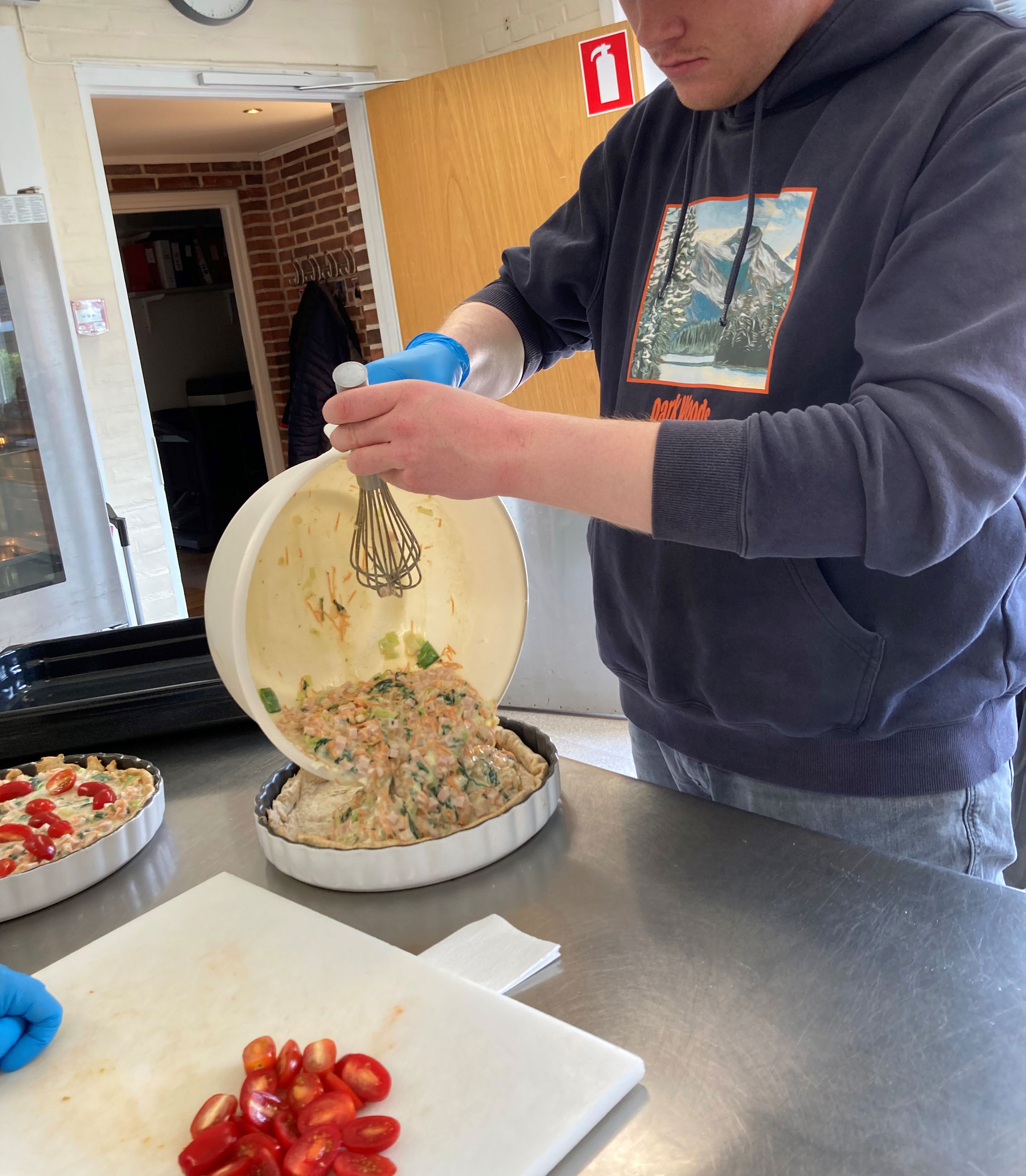 En borger står ved et tærtefad og hælder fyld af kød æg og porre ned i, ved siden af ham på skærebrættet ligger de halve tomater lige til at ligge ovenpå.