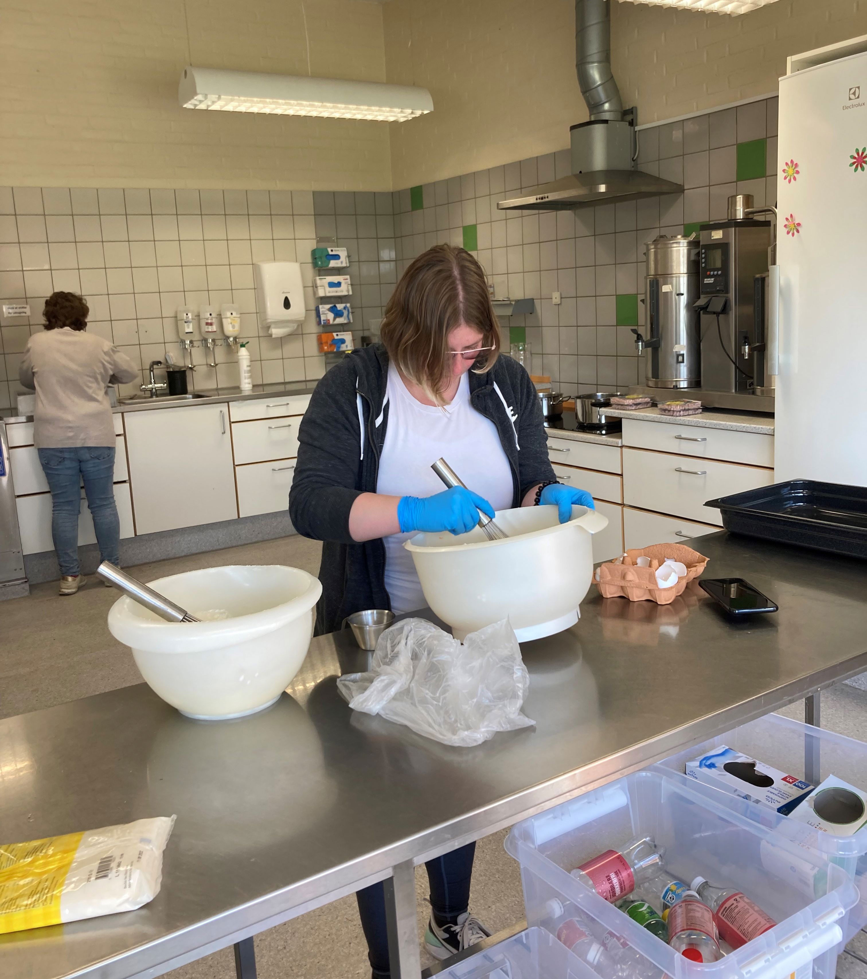 En borger står ved et stort bord og pisker æg sammen i stor skål, bag hende ser man en anden borger står ved vasken, på væggen på den anden side hænger der handsker i alle størrelser.