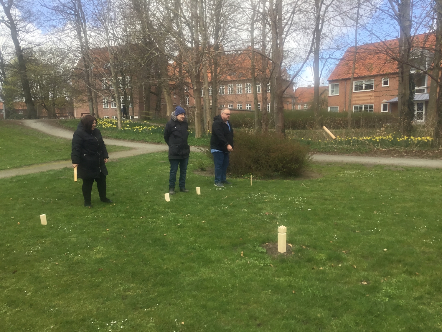 Tre borgere spiller kongespil på en grøn græsplæne, bag nogle store huse med røde mursten.