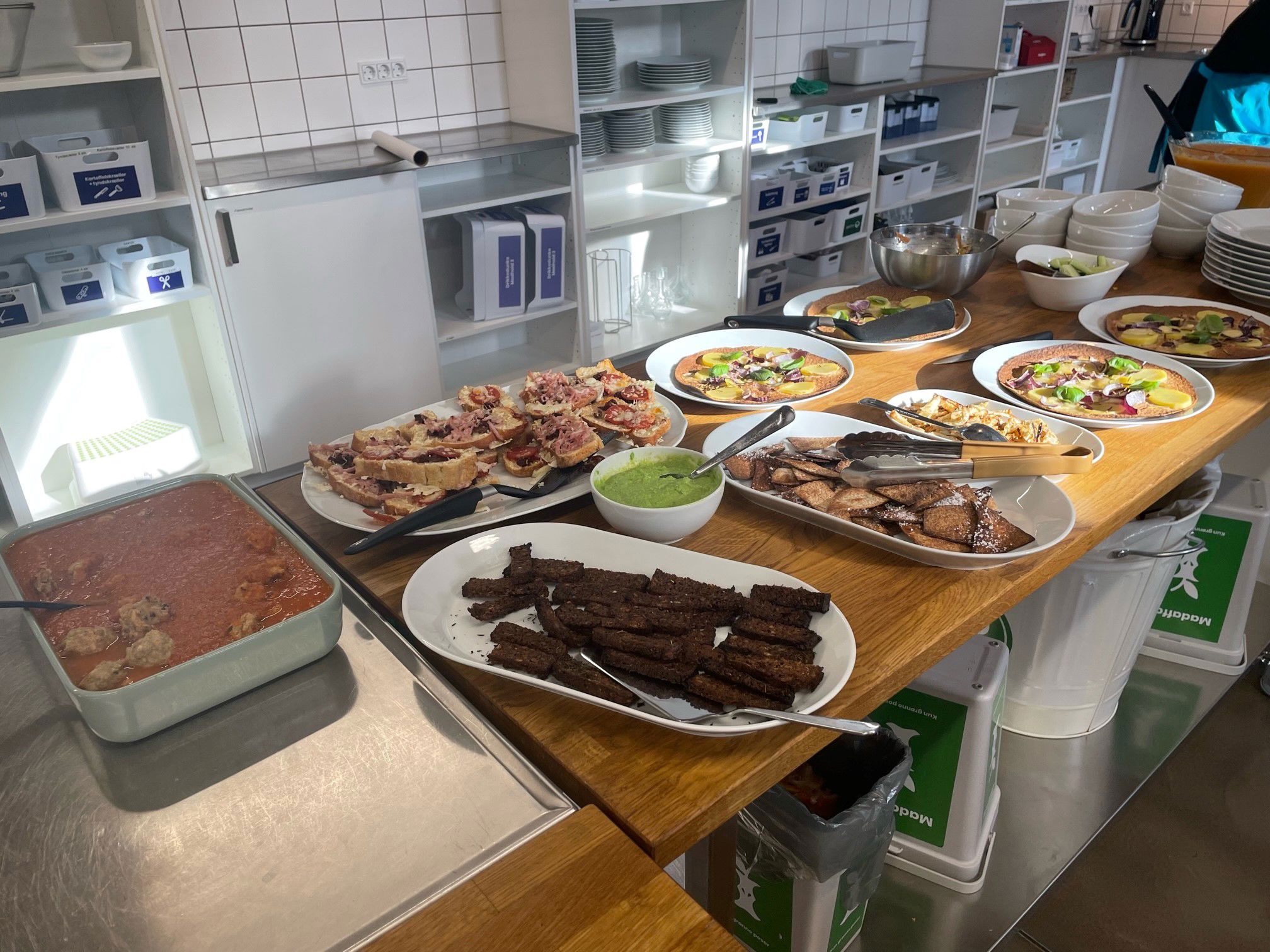 Køkkenbordplade i træ, på pladen står der en masse mad anrettet på forskellige hvide fade. På væggen bagved står en masse rent service.