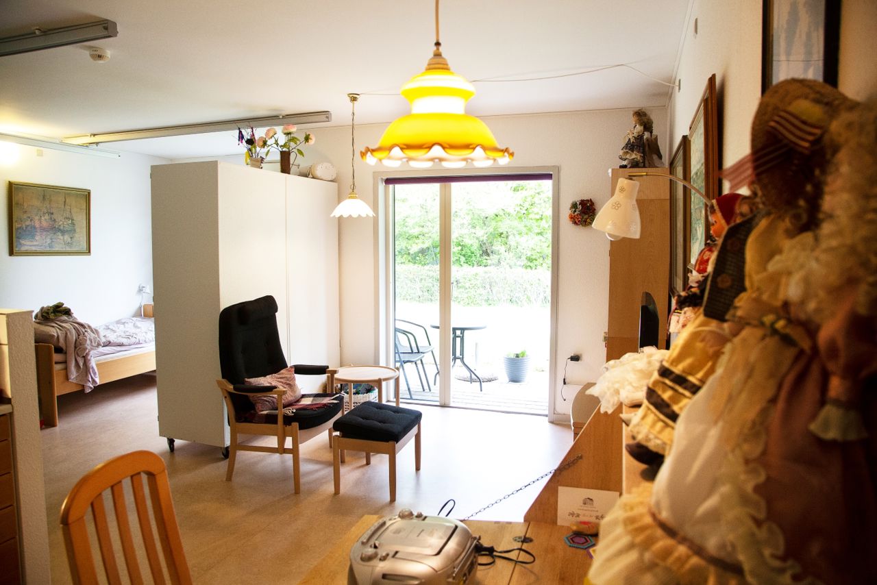 I foran ser man en skrivebord hvor der står nogle dukker på og i baggrunden står en seng og lænestol med skammel.