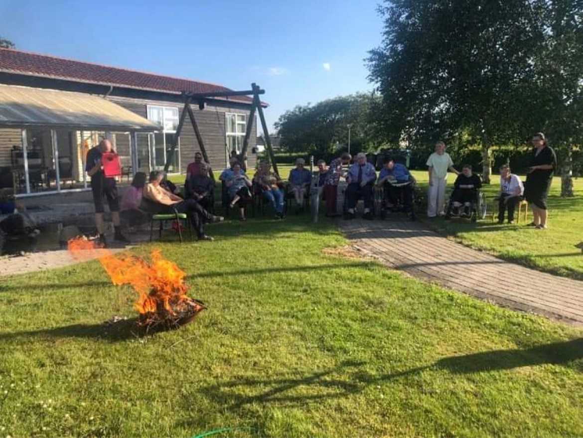 Tændt bål på græsplæne bagved er der flere borgere der kigger på bålet