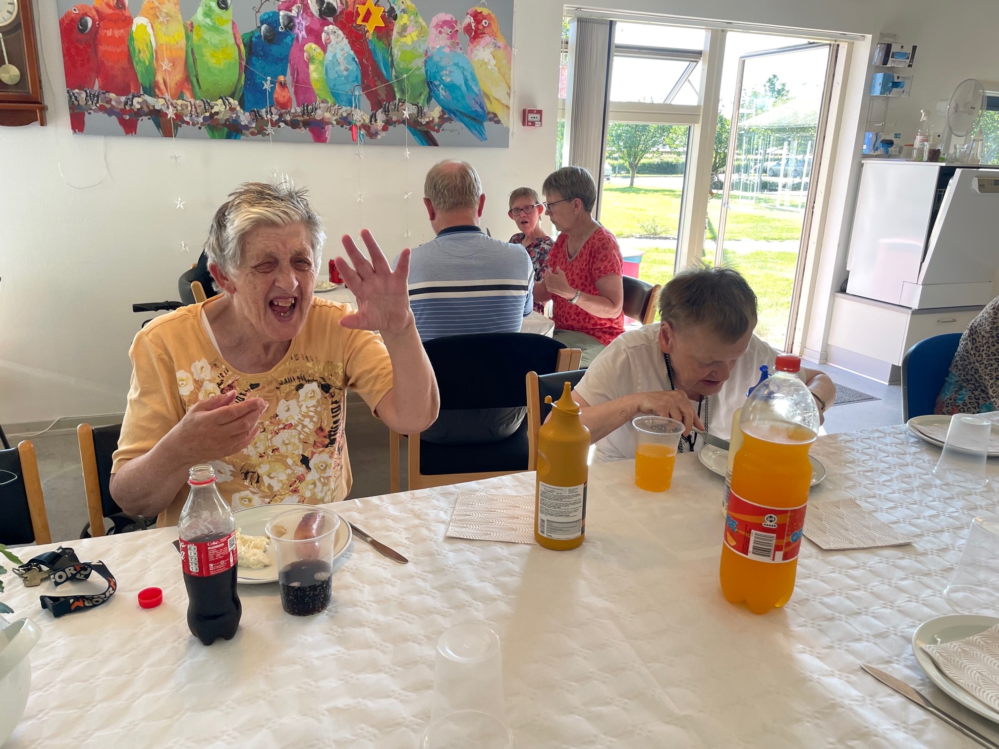 Borgerne sidder ved et bord og spiser gril mad, der er en hvid papirdug på bordene, på væggen bagved er et farverigt billede med papegøjer 