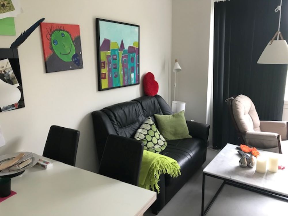 Stue hvor man forrest i billede kan se et lille hvidt spisebord med en sort stol til. Bagved det står der en sort lædersofa hvori de ligger et grønt tæppe og to grønne puder. Foran sofaen står der et lille marmor bord med en blomst og to bloklys.