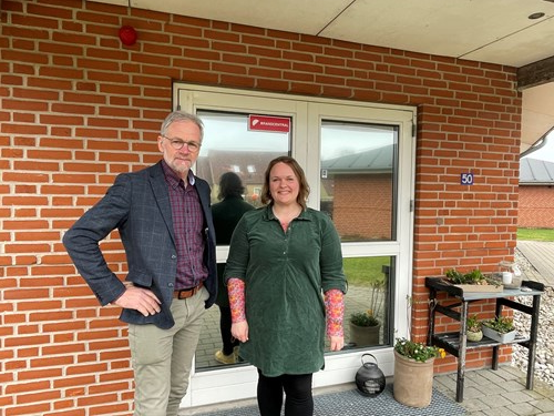 Borgmester og en medarbejder står foran en indgang, der står en blomsterkrukke ved døren og et lille bord hvor der også er blomster.