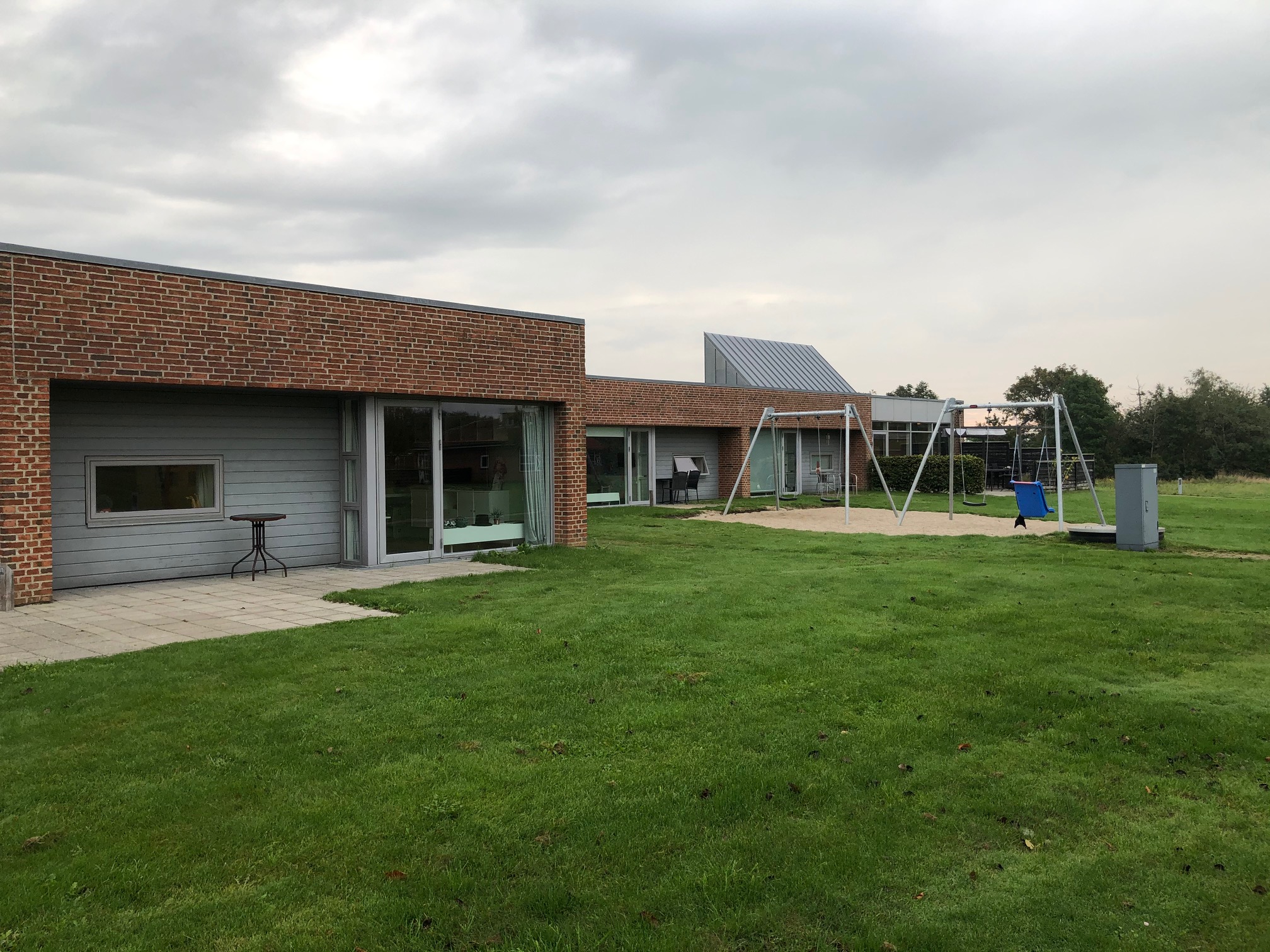 Æblevej udenfor, bygningerne er af røde mursten, og træ der er malet gråt. Foran bygninger er der en grøn græsplæne, og til højre i billedet er der to gyngestativer med hver to gynger på. Gyngestativerne er placeret ovenpå et område med sand.