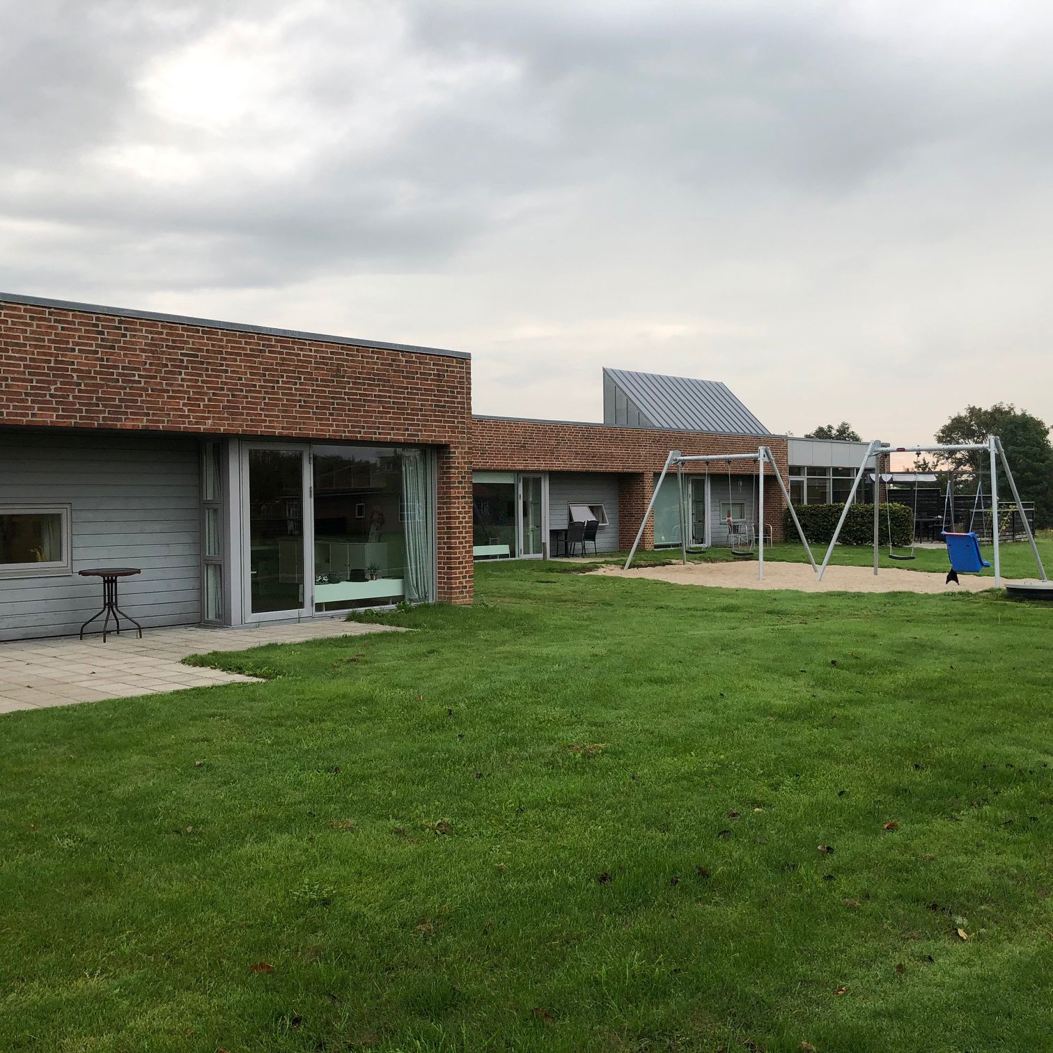 Æblevej udenfor, bygningerne er af røde mursten, og træ der er malet gråt. Foran bygninger er der en grøn græsplæne, og til højre i billedet er der to gyngestativer med hver to gynger på. Gyngestativerne er placeret ovenpå et område med sand.