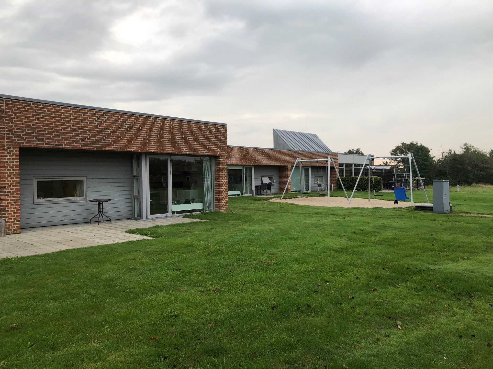 Æblevej udenfor, bygningerne er af røde mursten, og træ der er malet gråt. Foran bygninger er der en grøn græsplæne, og til højre i billedet er der to gyngestativer med hver to gynger på. Gyngestativerne er placeret ovenpå et område med sand.