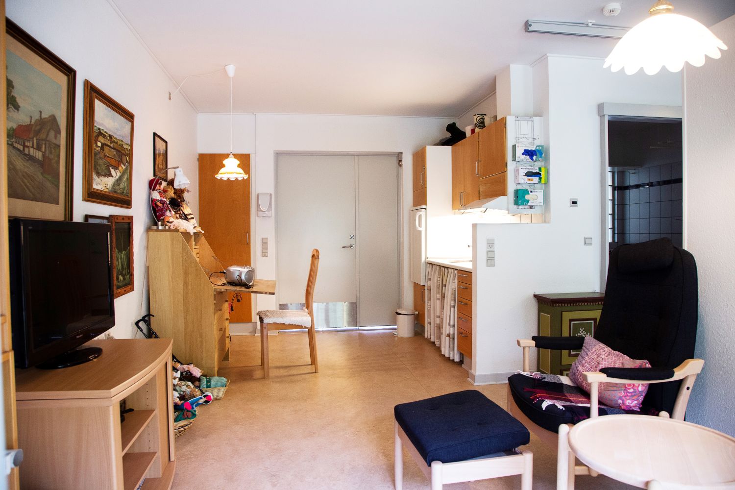 Foran i billedet står der en lænestol med skammel og kigger over på et fjernsyn som står på en lille reol. På vægen hænger der billeder og i baggrunden er der en lille skrivebord med stol og bagved et lille tekøkken.