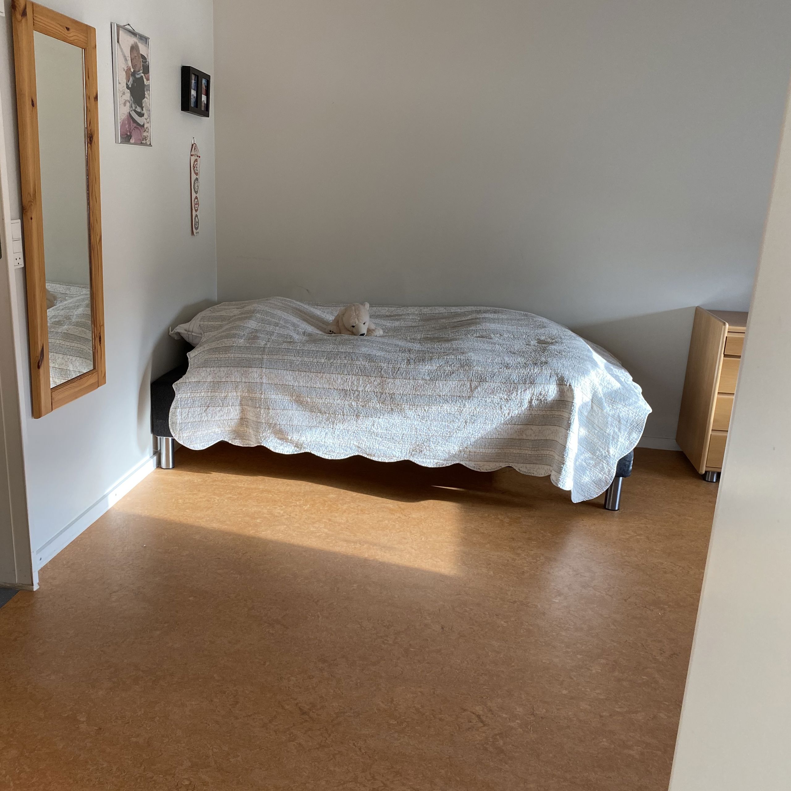 En borgers lejlighed hvor der er en seng. Gulvet er i lysebrune farver. Der hænget et langt spejl på væggen ind til toilettet.