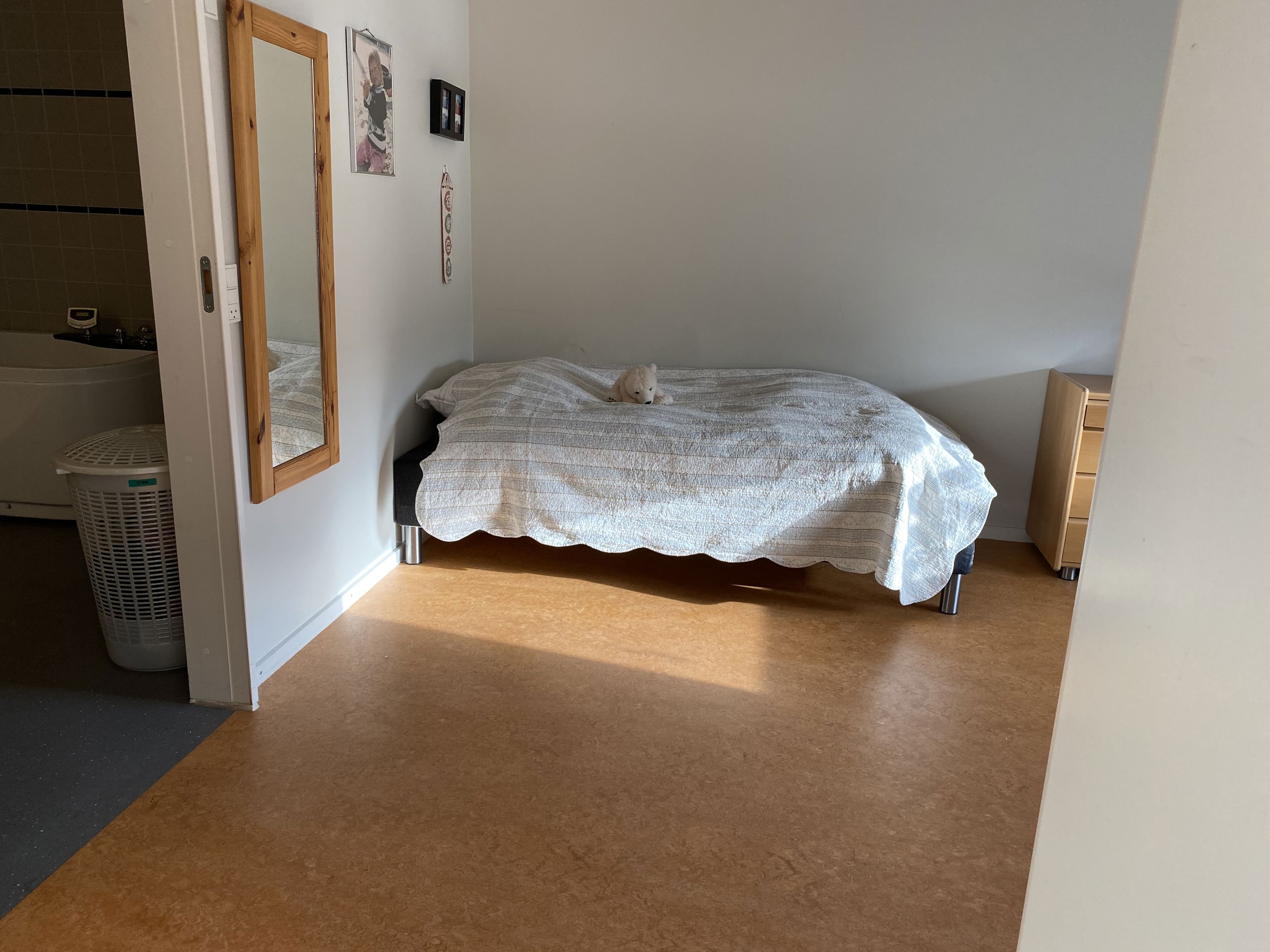 En borgers lejlighed hvor der er en seng. Gulvet er i lysebrune farver. Der hænget et langt spejl på væggen ind til toilettet.