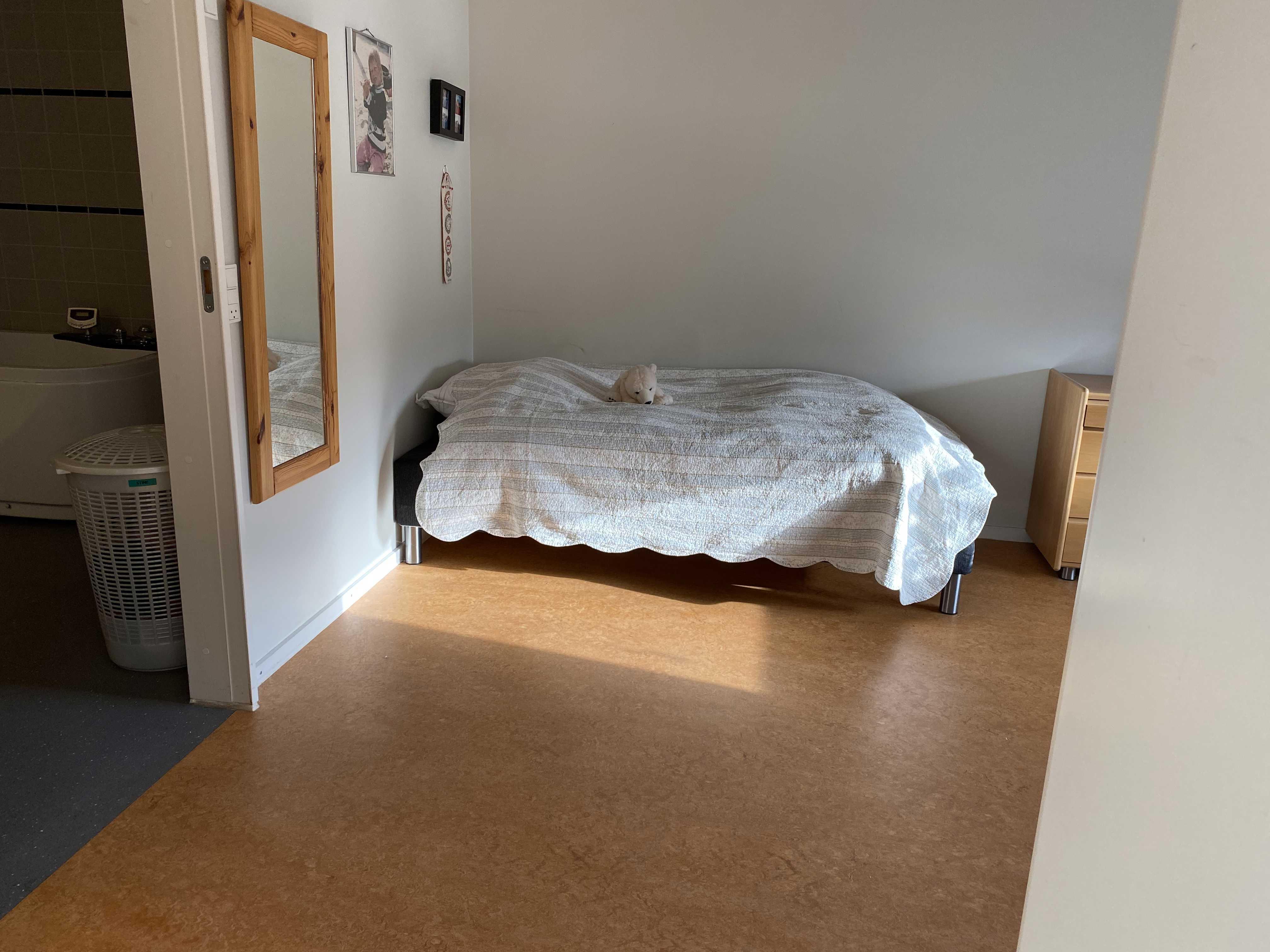 En borgers lejlighed hvor der er en seng. Gulvet er i lysebrune farver. Der hænget et langt spejl på væggen ind til toilettet.