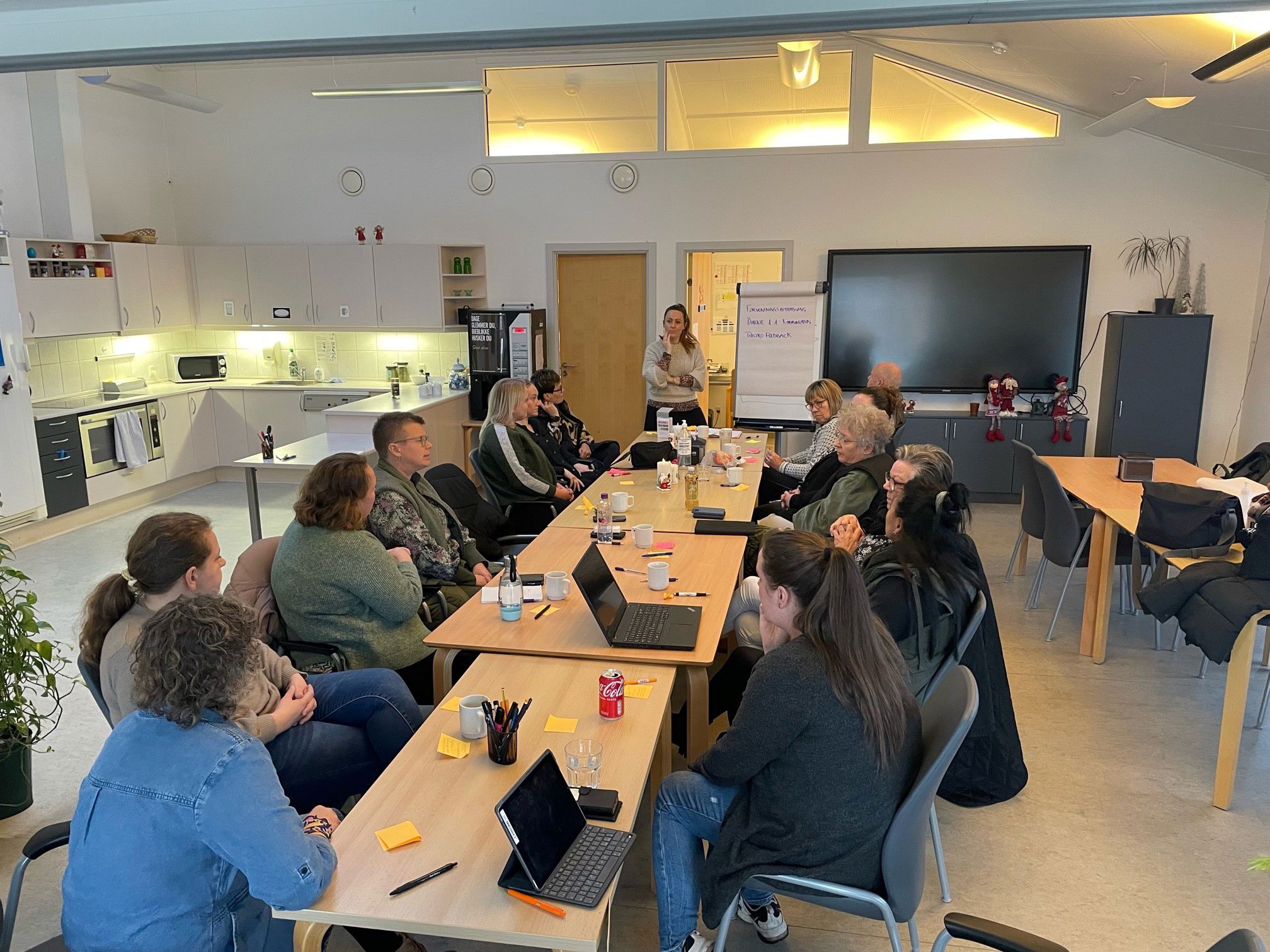 Et langt bord hvor der sidder personer og lytter til en foredragsholder, som står for enden af bordet. Bagerst i lokalet er en stor skærm og et skab, hvor der sidder nisser.