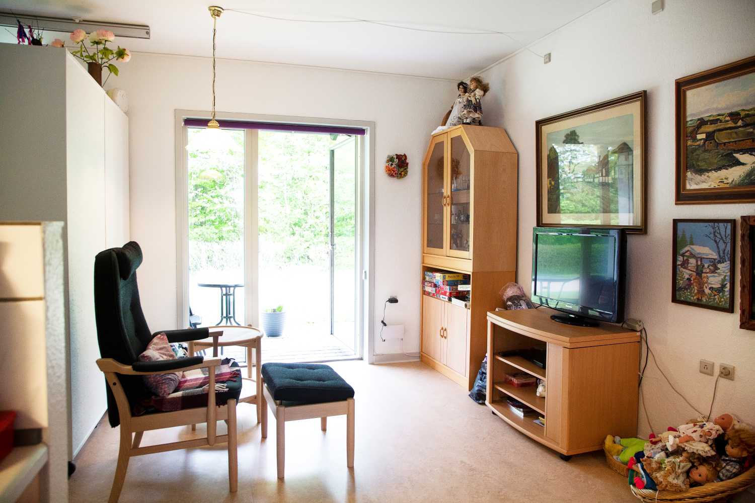 Foran i billedet står der en lænestol med skammel og kigger over på et fjernsyn som står på en lille reol. Og i baggrunden et stort vindue hvor man kigger ud på haven.