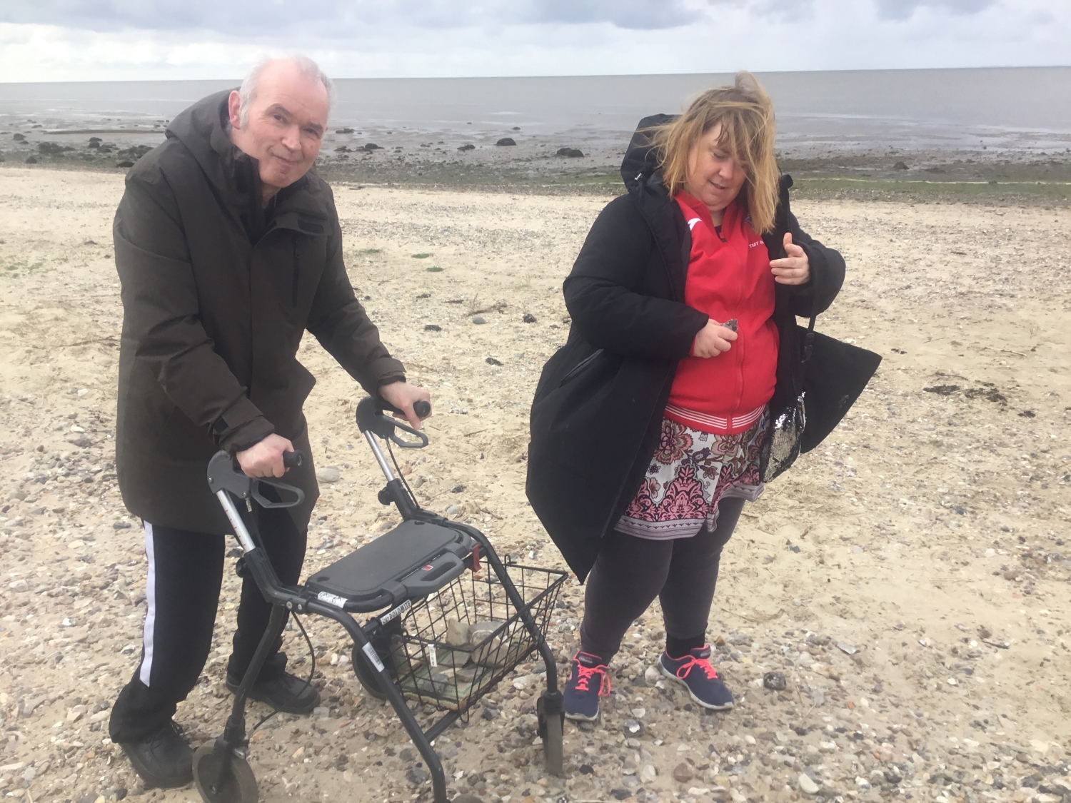 To borgere står på stranden i det gule sand med vandet i baggrunden, den ene har en rollator med, de ser begge glade ud.
