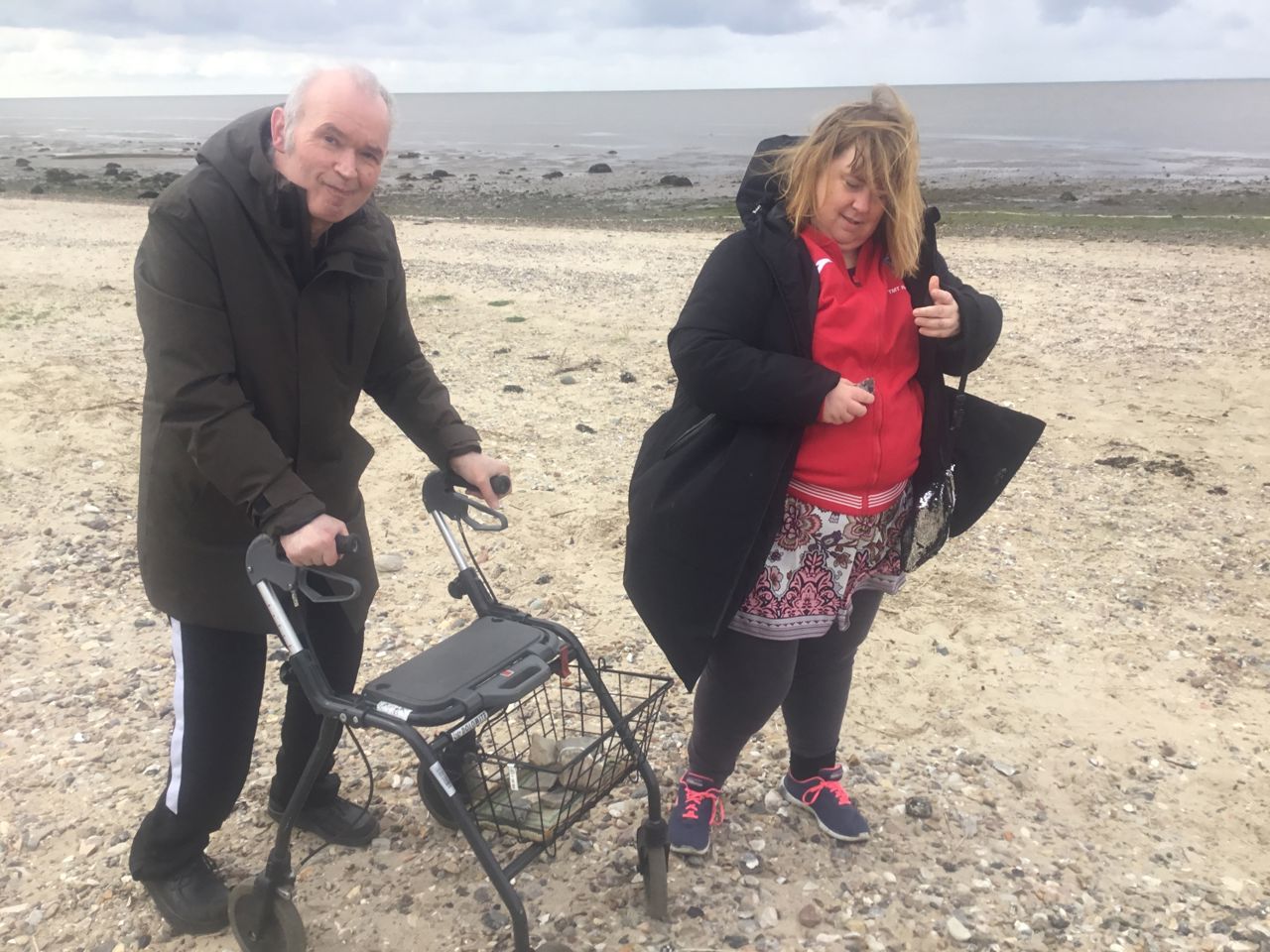 To borgere står på stranden i det gule sand med vandet i baggrunden, den ene har en rollator med, de ser begge glade ud.
