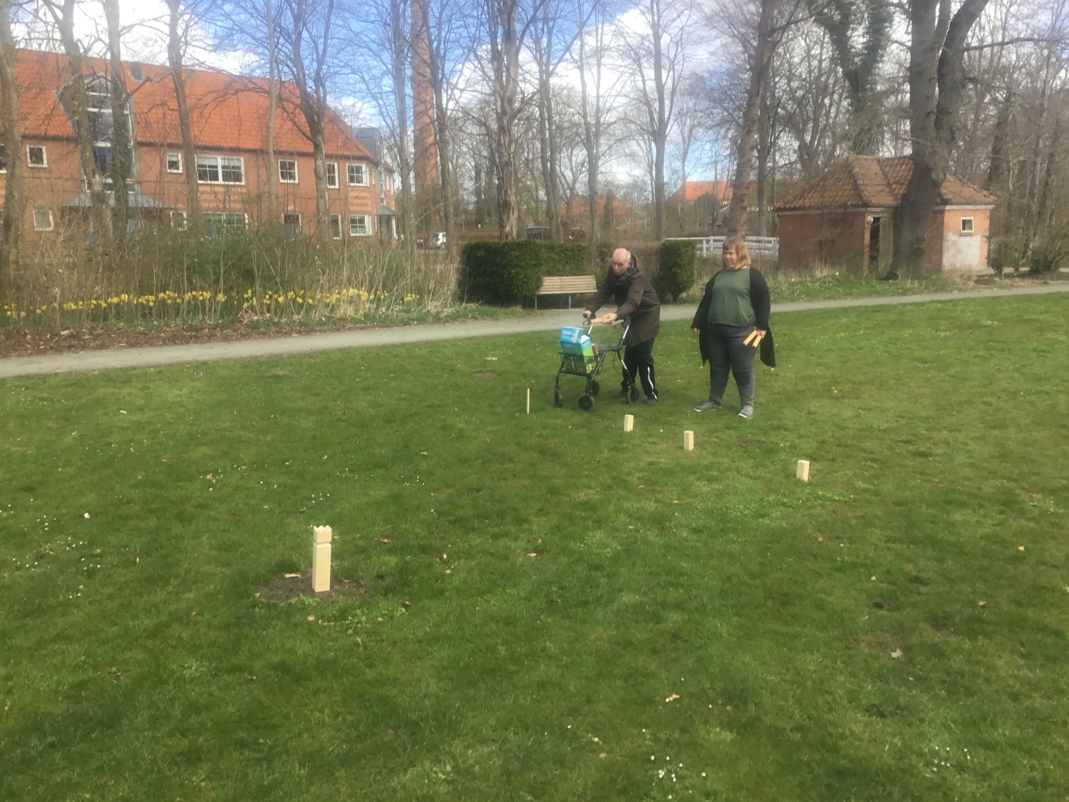 To borgere spiller kongespil på en grøn græsplæne, den ene borger har en rollator med, i baggrunden er der store huse med røde mursten