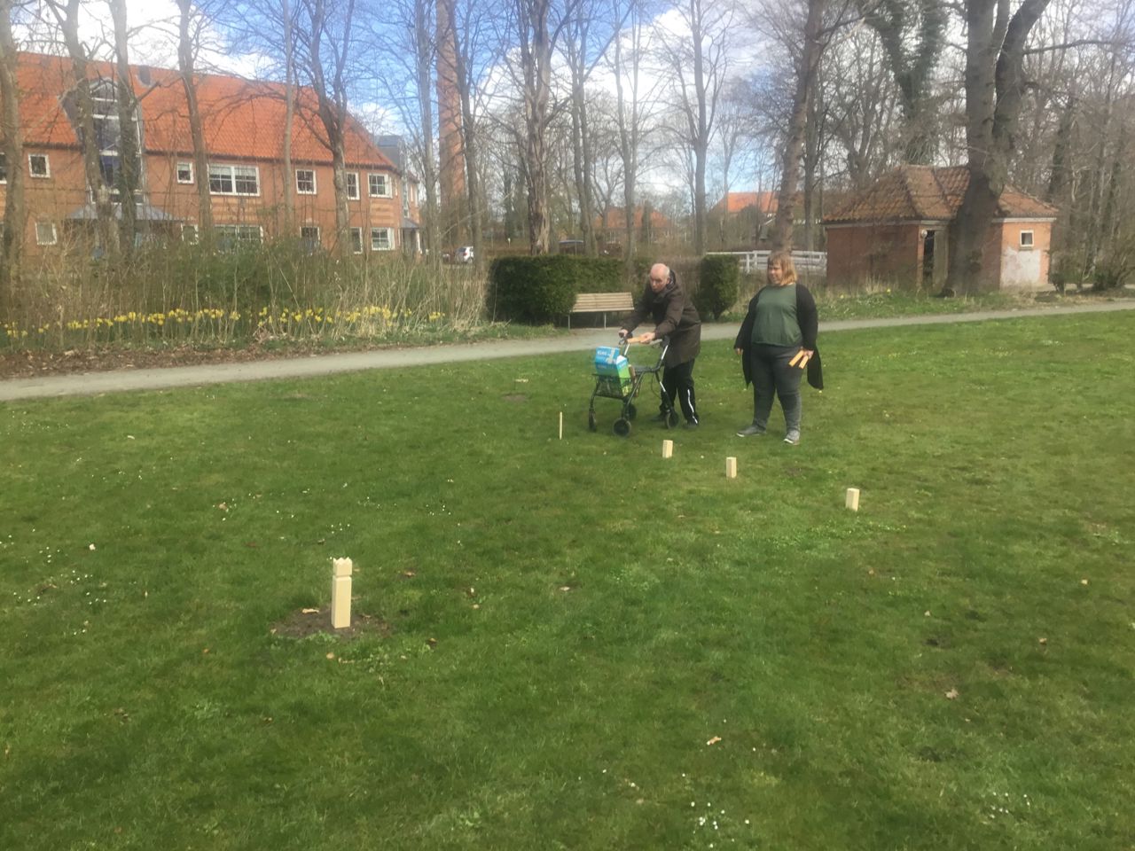 To borgere spiller kongespil på en grøn græsplæne, den ene borger har en rollator med, i baggrunden er der store huse med røde mursten
