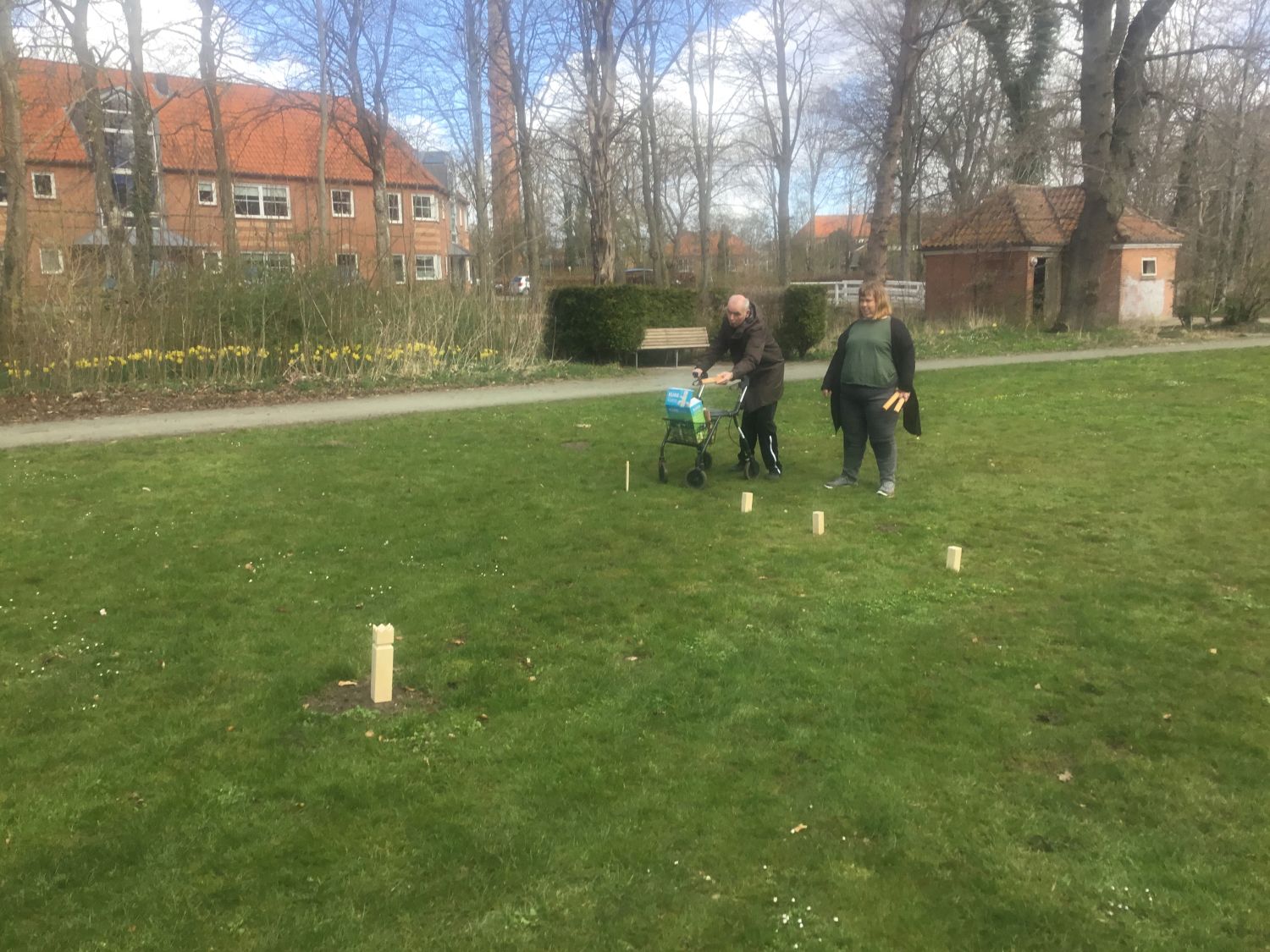 To borgere spiller kongespil på en grøn græsplæne, den ene borger har en rollator med, i baggrunden er der store huse med røde mursten