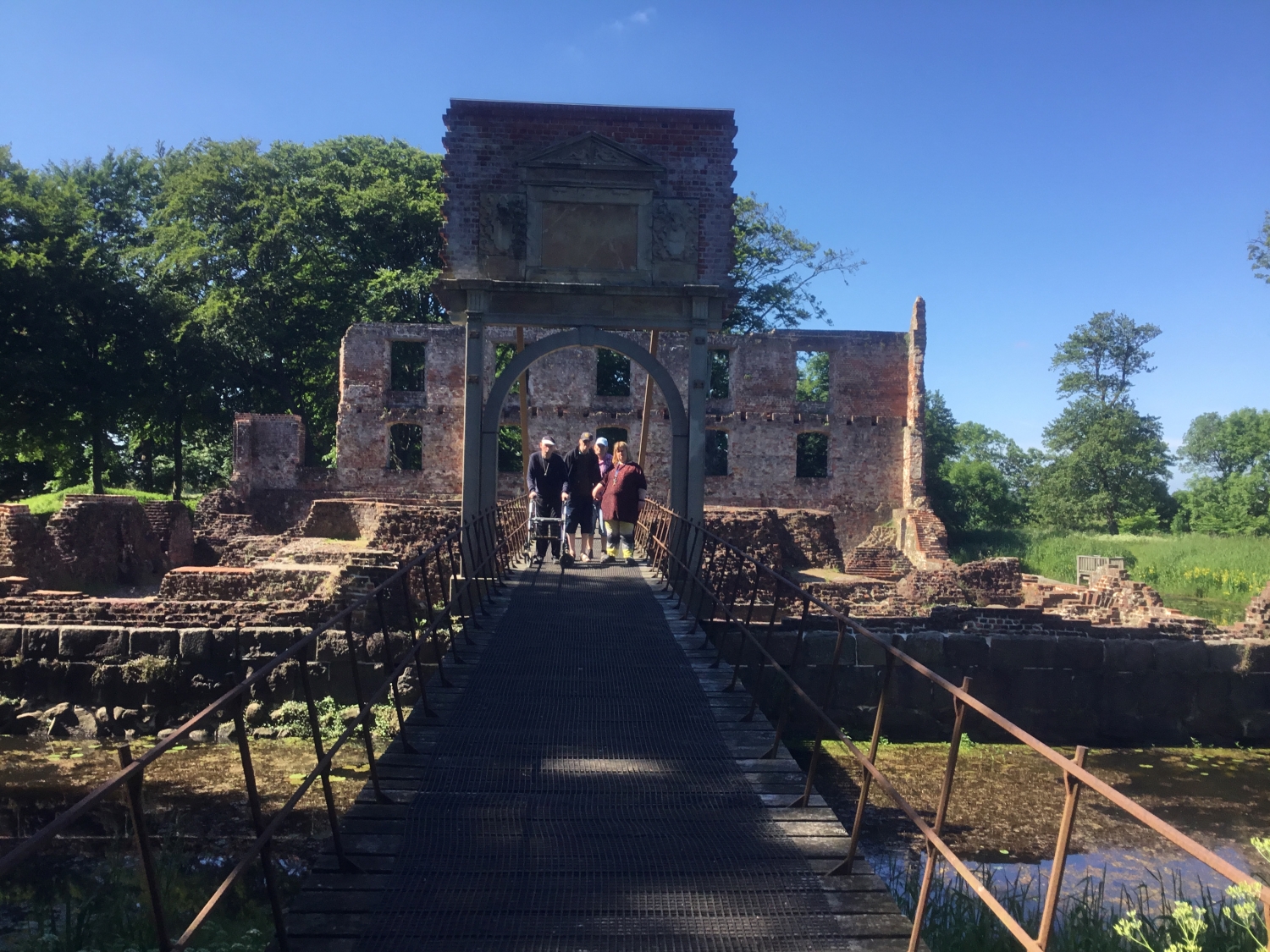 Fire borgere på en metalbro over til en ruin, den er i røde mursten, man kan se vinduer og foran er der vand som broen går henover