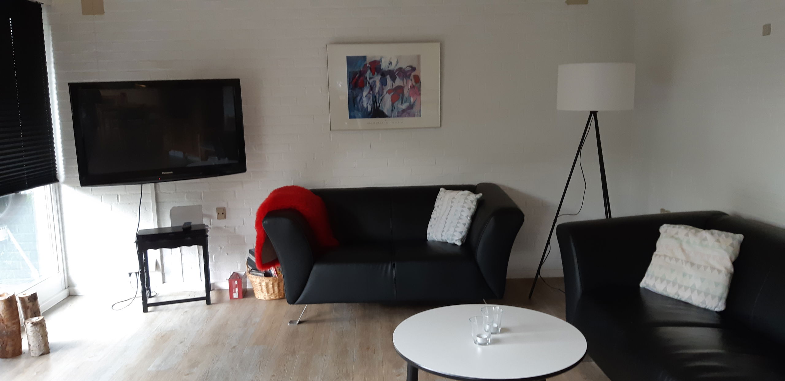 Opholdsstue med to lædersofaer, et rundt bord og et tv. I hjærnet er der er høj standerlampe med hvid lampeskærm.