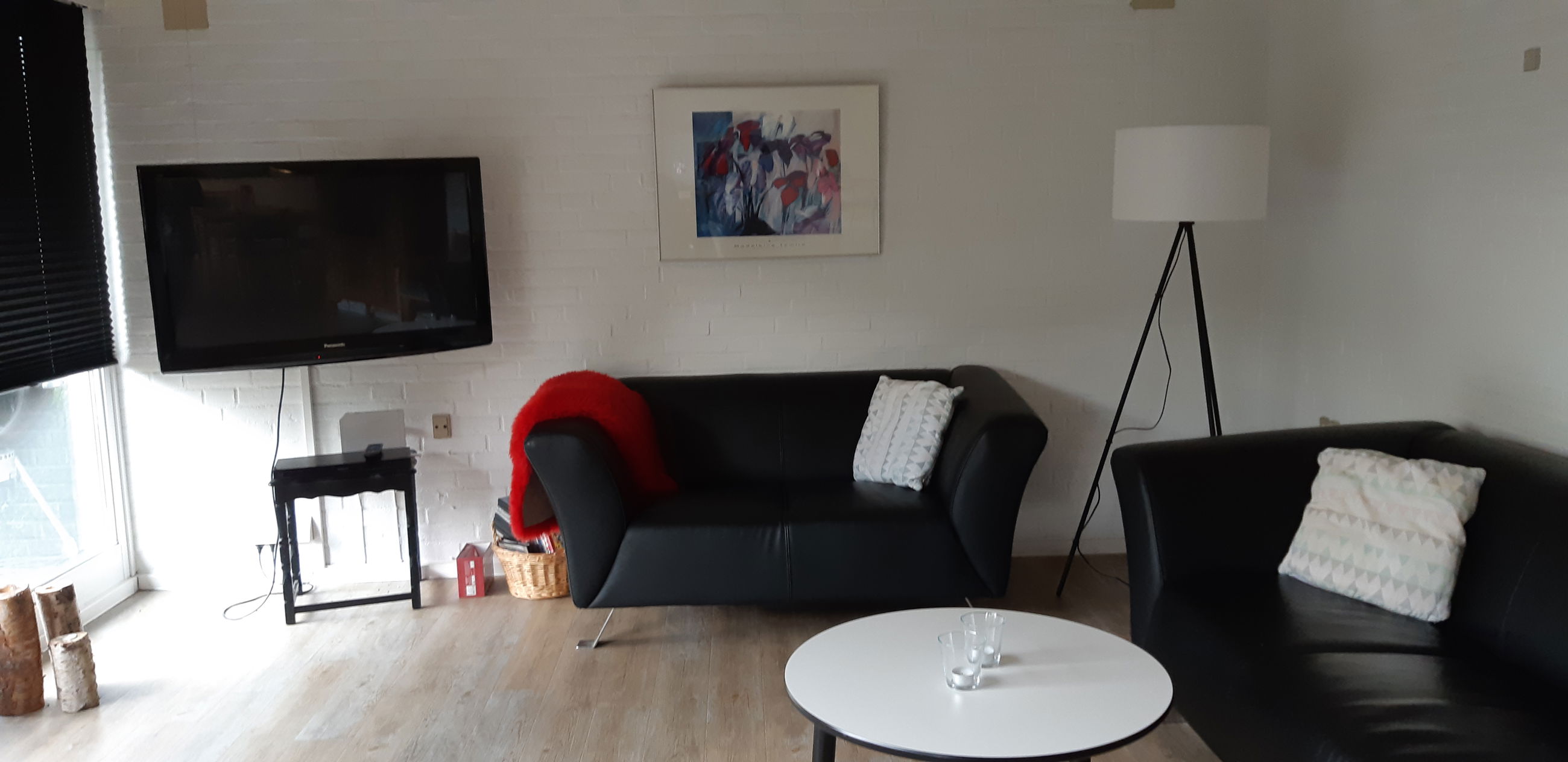 Opholdsstue med to lædersofaer, et rundt bord og et tv. I hjærnet er der er høj standerlampe med hvid lampeskærm.